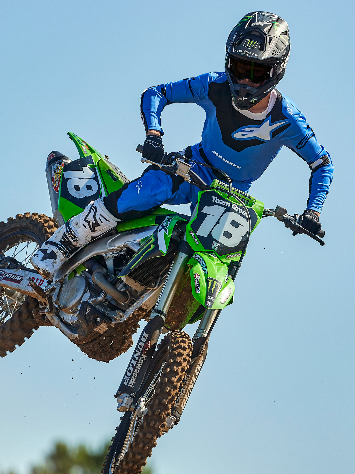 Alpinestars 2025 Racer Veil Jersey, motocross trøje, blå og sort, langærmet teknisk top med abstrakte grafiske mønstre og åndbare mesh-sektioner, motocross-kører i luften på en grøn motorcykel, professionelt off-road racerudstyr