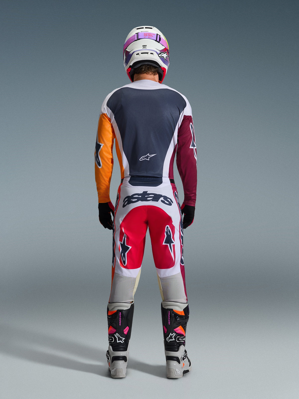 Motocrosskører set bagfra iført komplet racerudstyr, 2026 Racer Portl Jersey i rød, lysegrå og mørkegrøn med asymmetriske orange og bordeauxrøde ærmer, matchende hvide og røde bukser, hvid hjelm med lilla goggles, hvide og sorte støvler, koordineret sæt til off-road kørsel