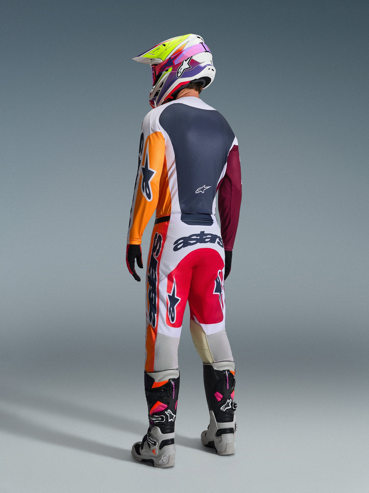Motocross-kører set bagfra iført komplet racerudstyr, Alpinestars 2026 Racer Portl-trøje i rød, lysegrå og mørkegrøn med orange og bordeauxrøde paneler, matchende hvide og røde bukser, grå og sorte støvler med lyserøde detaljer, flerfarvet hjelm, koordineret off-road raceroutfit.