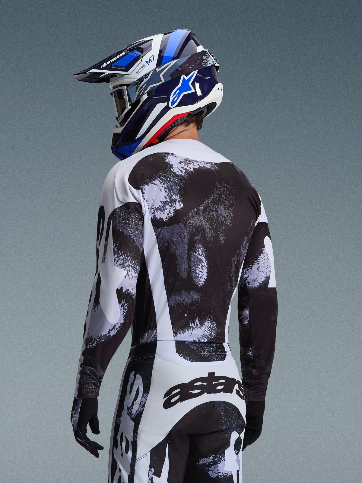 Alpinestars 2026 Racer Lahnd Jersey i Iron Camo, set bagfra af en motocrosskører i et sort, hvidt og gråt udstyrssæt med abstrakt mønster, inklusiv matchende bukser, sorte handsker og en blå racerhjelm med crossbriller.