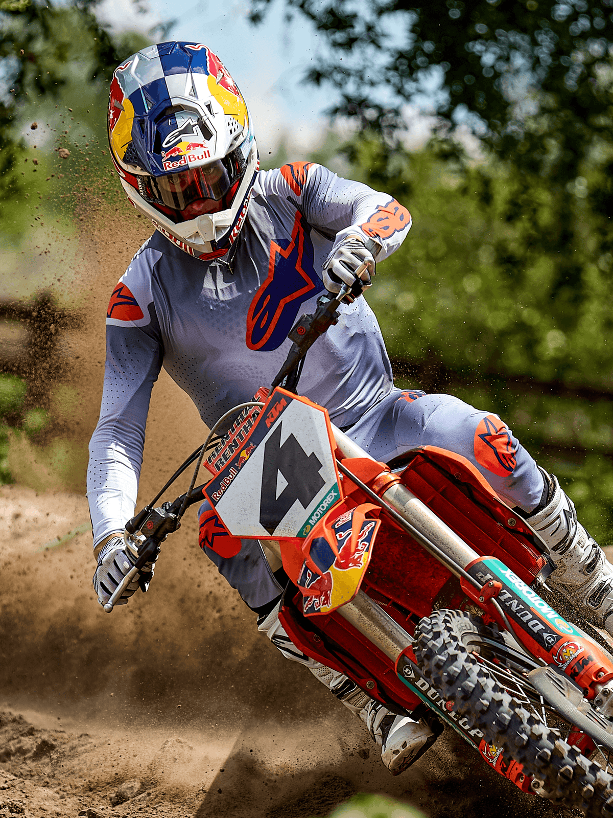 Motocrosskører i aktion på en jordbane iført Alpinestars 2025 Supertech Lipan Jersey i Gray Orange Fluo, matchende grå bukser med orange detaljer, hvide støvler og en Red Bull-hjelm, tager sving på en orange motocrosscykel med jordstænk, professionelt off-road racerudstyr