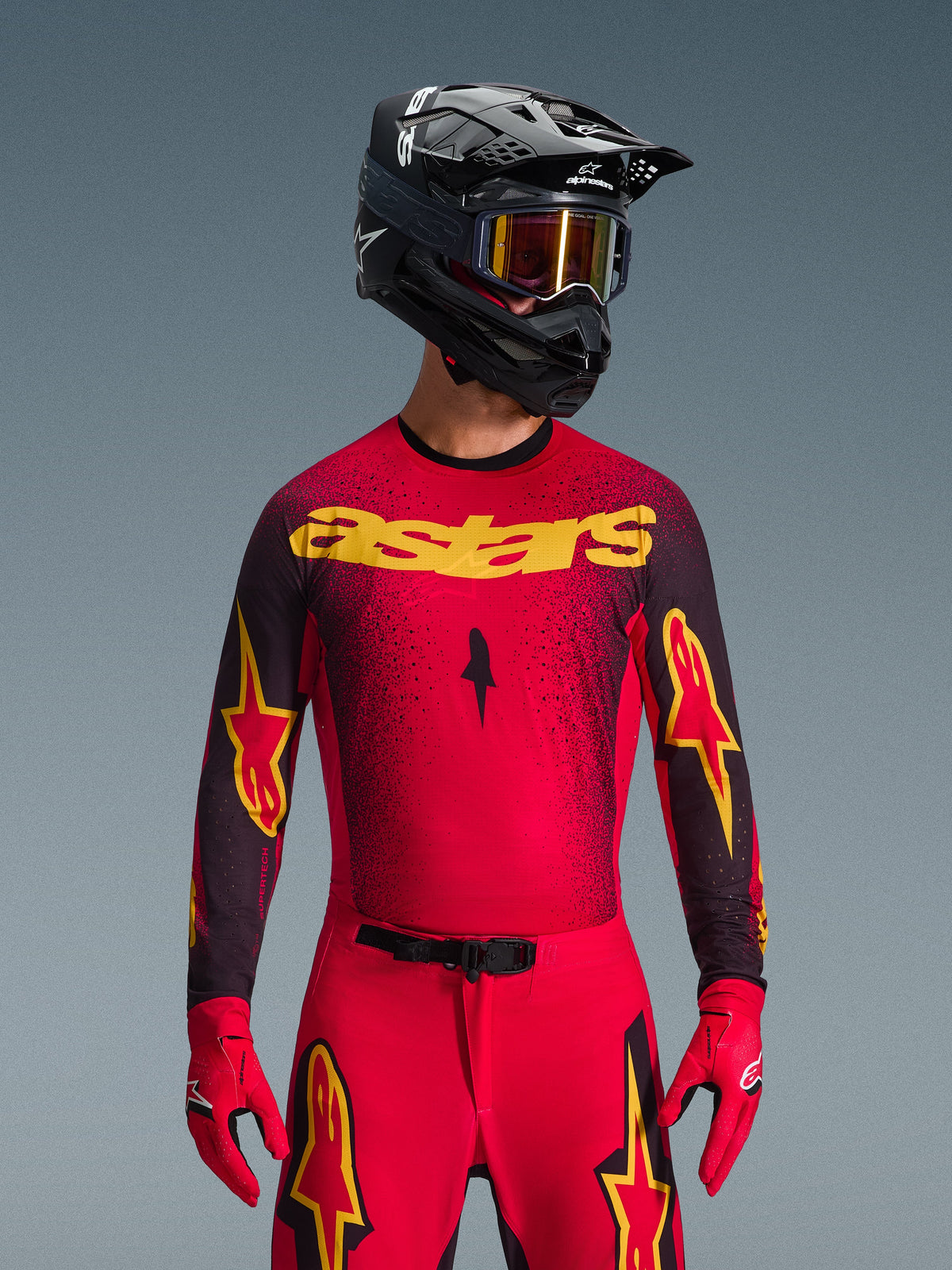 Alpinestars 2026 Supertech Scenz Gear Set, motocross-kører iført rød trøje med gul 'astars'-branding og sorte nistrede detaljer, røde bukser med gule stjernelogoer på lårene, røde racinghandsker og sort hjelm med reflekterende gule briller, professionelt raceroutfit til terrænkørsel