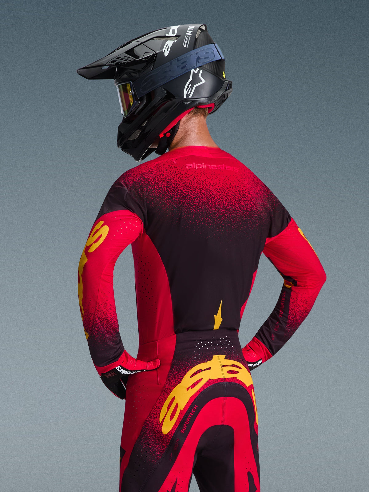 Alpinestars 2026 Supertech Scenz-trøje og matchende bukser, motocrosskører set bagfra, rød, sort og gul farvekombination, sort racerhjelm med hvidt logo, markant gul branding på ærmerne og bagpå, professionelt terrænkørselsudstyr