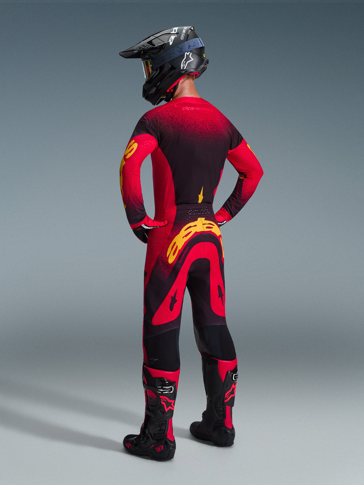 Motocrosskører iført Alpinestars 2026 Supertech Scenz-udstyrssæt, rød og sort trøje med gule detaljer, matchende bukser med markant gult logo på bagsiden, sort hjelm og støvler med røde detaljer, koordineret outfit til professionel motocross-racing