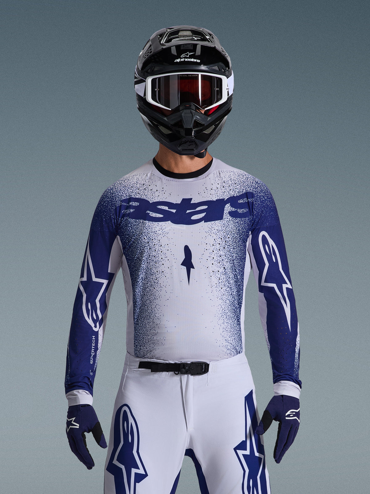 Motocross-kører iført Alpinestars 2026 Supertech Scenz-udstyrssæt, hvid jersey med en blåprikket graduering og 'astars'-branding, matchende hvide bukser med blå detaljer, sort hjelm med hvide goggles og blå handsker, som viser den atletiske professionelle racerpasform.