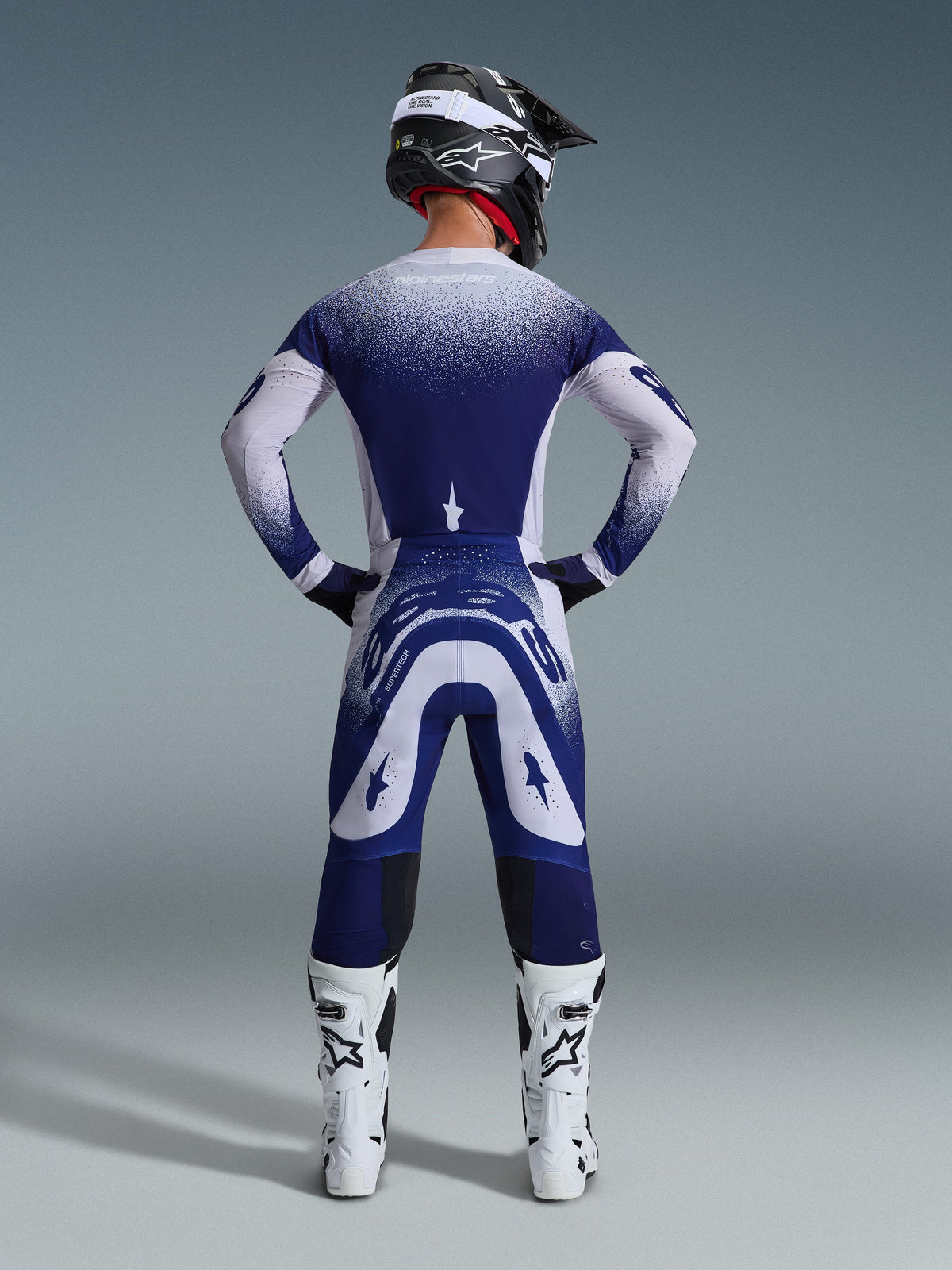 Alpinestars 2026 Supertech Scenz-udstyrssæt, set bagfra af en motocross-kører i blå og hvid racing-jersey og bukser, prikket gradienteffekt, båret med hvide motocrossstøvler og sort hjelm, professionelt udstyr til terrænkørsel
