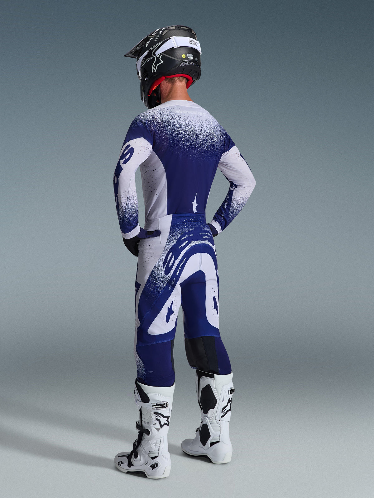 Motocross-kører iført Alpinestars 2026 Supertech Scenz-udstyrssæt, marineblå og hvid jersey og bukser med en spættet gradienteffekt, set bagfra med atletisk pasform, kombineret med hvide og sorte støvler og en sort hjelm, professionelt racerudstyr til terrænkørsel