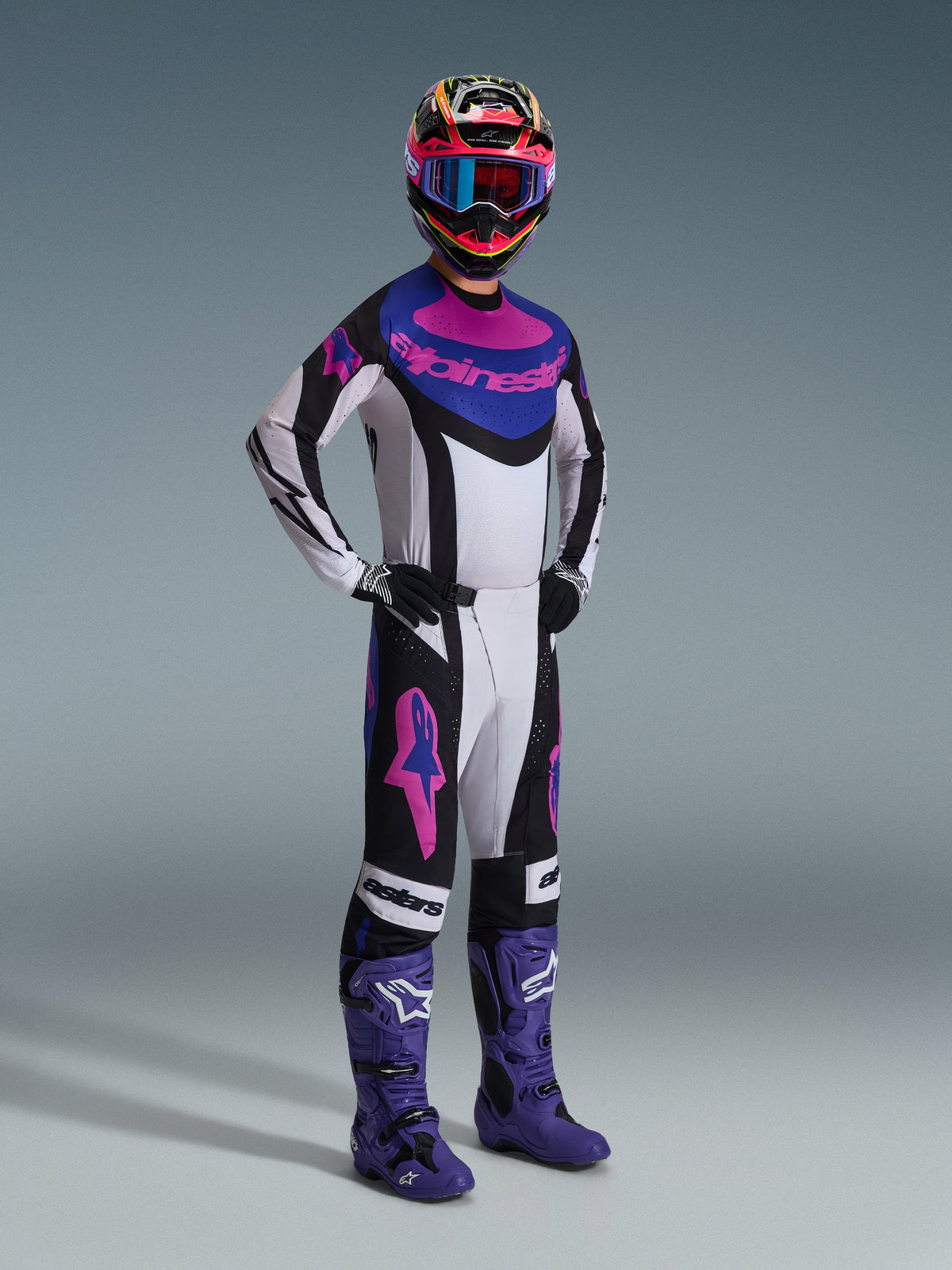 Motocross-kører iført Alpinestars 2026 Techstar Knif Jersey i Sort Lysgrå Lilla, matchende bukser med store lyserøde logoer, lilla støvler og en flerfarvet hjelm med blå goggles, komplet racersæt til off-road ydeevne