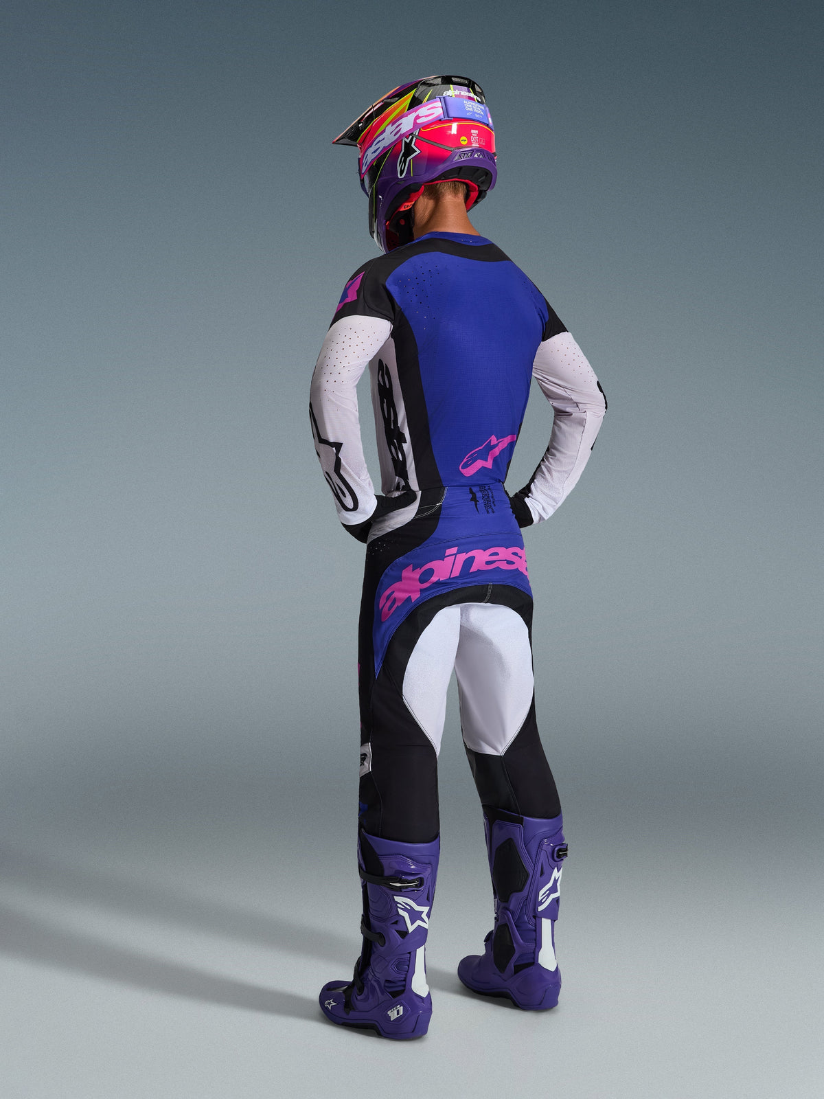 Motocrosskører set bagfra iført komplet racerudstyr, 2026 Techstar Knif Jersey med blå ryg og hvide perforerede ærmer, matchende sorte bukser med pink Alpinestars-branding, farverig hjelm og lilla støvler, koordineret outfit til off-road konkurrence