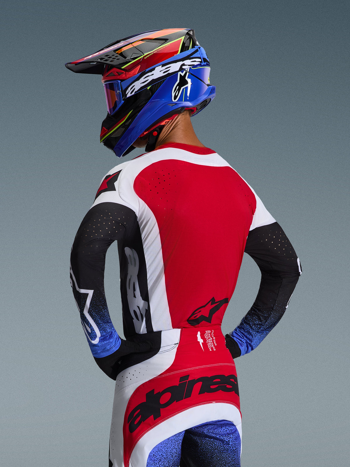 Alpinestars 2026 Techstar Knif-udstyrssæt, motocross-rytter set bagfra, rød trøje med hvide sidepaneler, sorte ærmer med blåspættede manchetter, røde og hvide bukser med sort Alpinestars-branding over sædet, matchende blå og rød hjelm med goggles