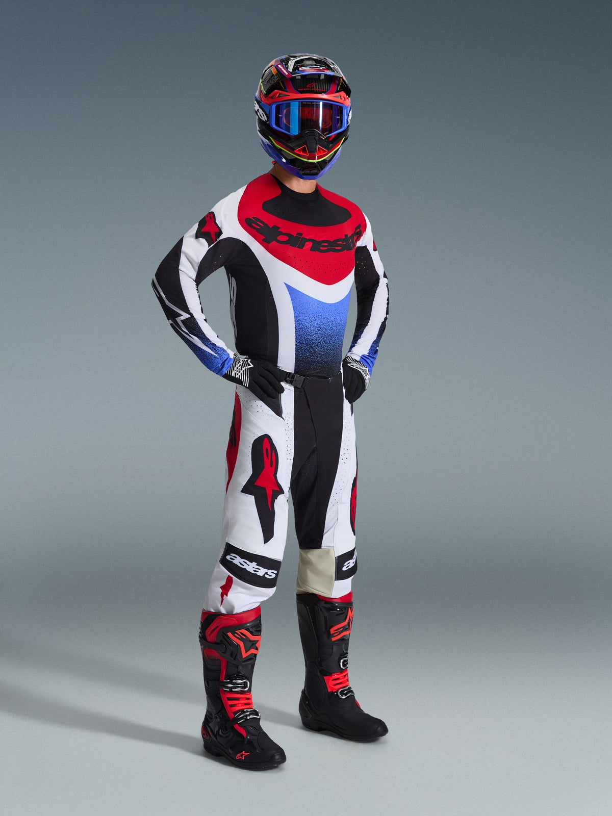Alpinestars 2026 Techstar Knif-udstyrssæt, motocross-kører med trøje i sort, hvid og rød med blåt farveforløb på brystet, hvide bukser med røde logoer og sorte detaljer, sorte og røde støvler, professionelt racerudstyr til off-road