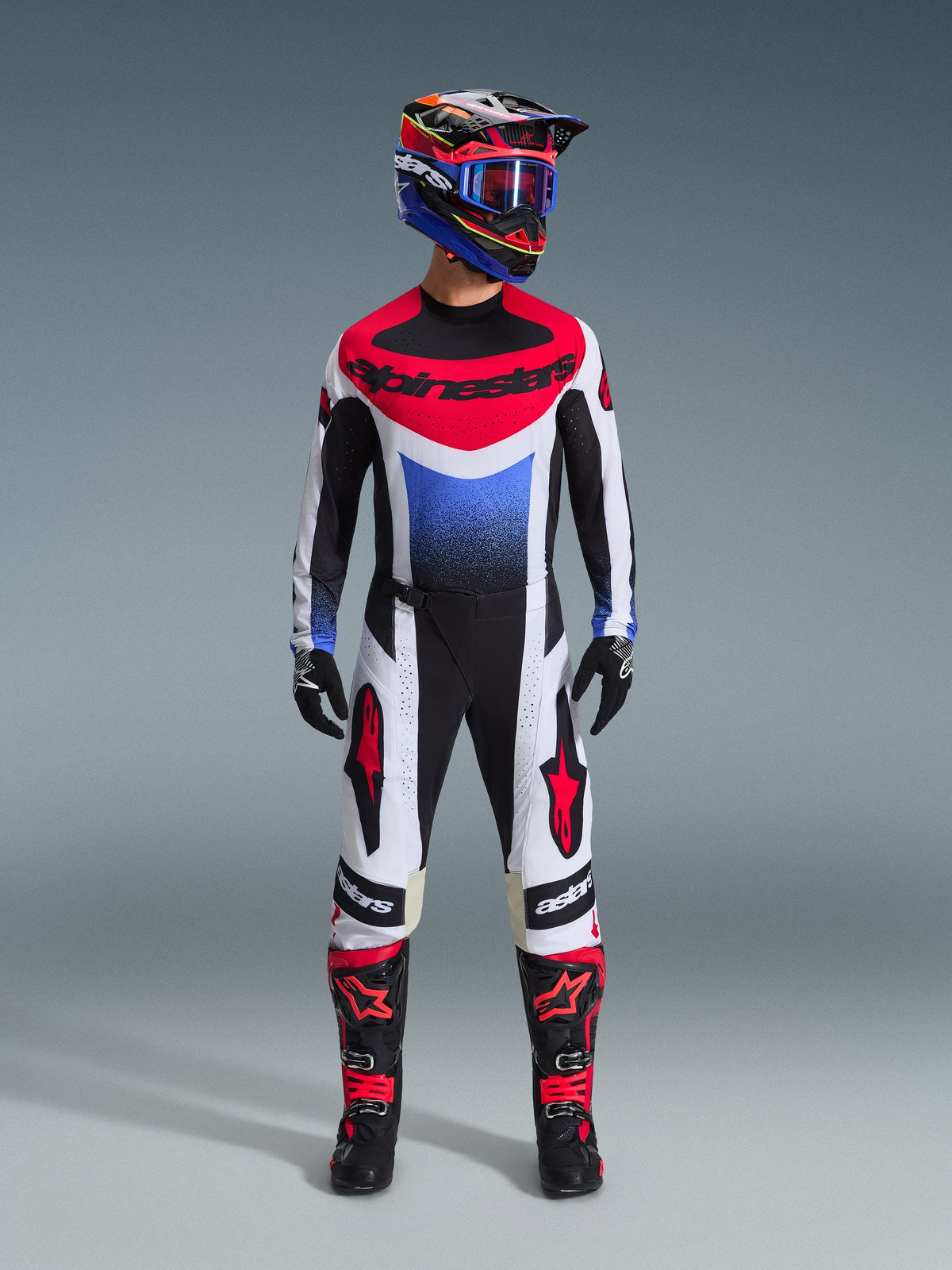 Motocross-kører iført Alpinestars 2026 Techstar Knif-udstyrssæt, sort, hvid og rød trøje med blå gradient og branding, matchende sorte og hvide bukser med røde detaljer, flerfarvet hjelm med blå goggles og sorte og røde støvler, koordineret off-road raceroutfit.