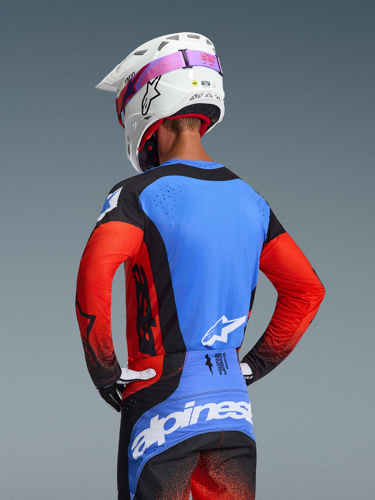 Alpinestars 2026 Techstar Knif Gear Set, motocross-udstyr, kører set bagfra, blå jersey med røde ærmer og sorte detaljer, sorte og blå bukser med hvidt logo, hvid hjelm med lyserød brillerem, laserskårne perforationer til ventilation