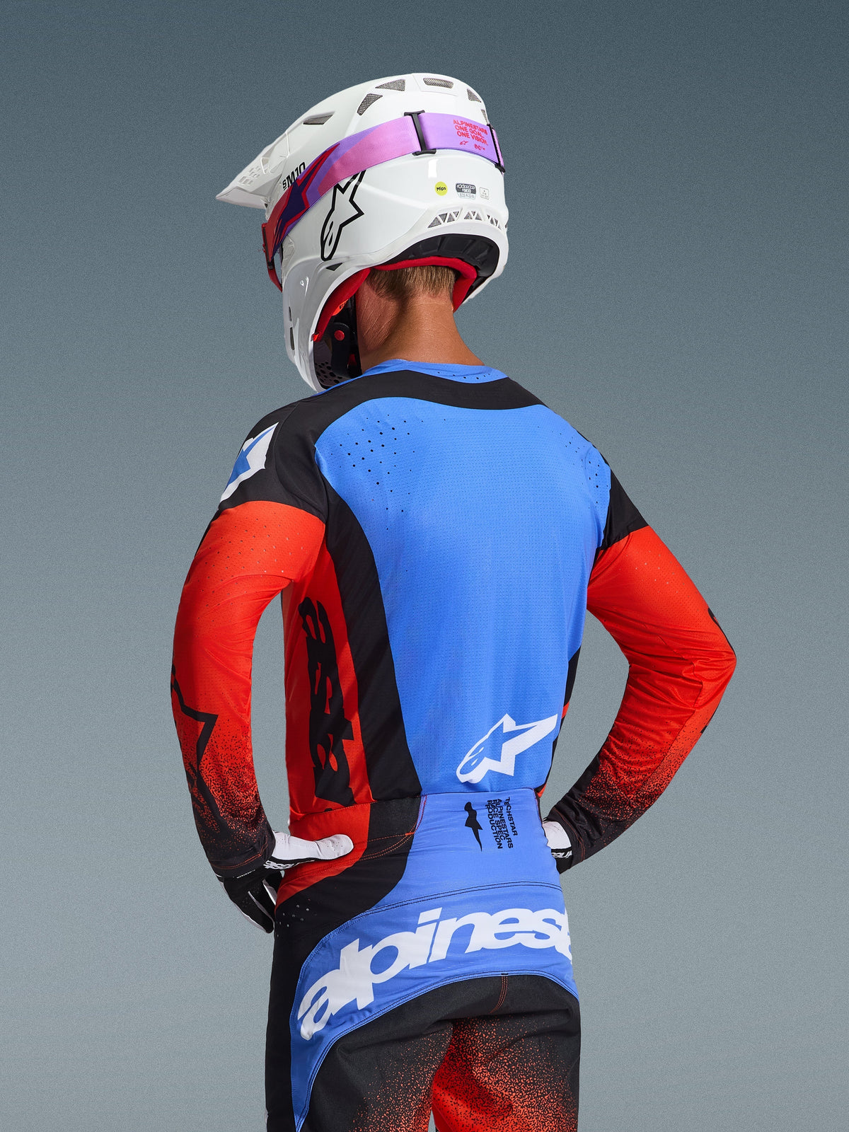 Alpinestars 2026 Techstar Knif Jersey, motocross-trøje set bagfra i Orange Black UCLA Blue, blåt rygpanel med laserskårne perforeringer, orange ærmer og sorte detaljer, matchende motocrossbukser og hvid Alpinestars-hjelm