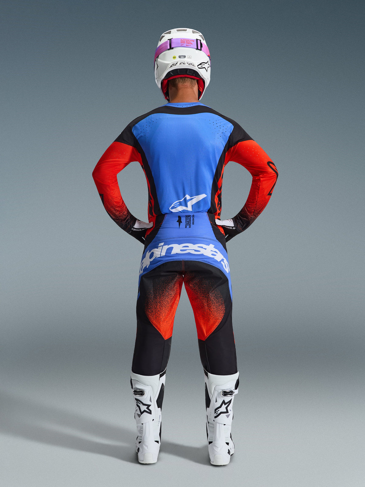 Bagfra-visning af en motocross-kører iført Alpinestars 2026 Techstar Knif-udstyrssæt, blå og rød trøje med sorte detaljer, sorte bukser med rød gradient på lårene og blå branding, kombineret med hvide racerstøvler og en hvid hjelm med pink goggles.