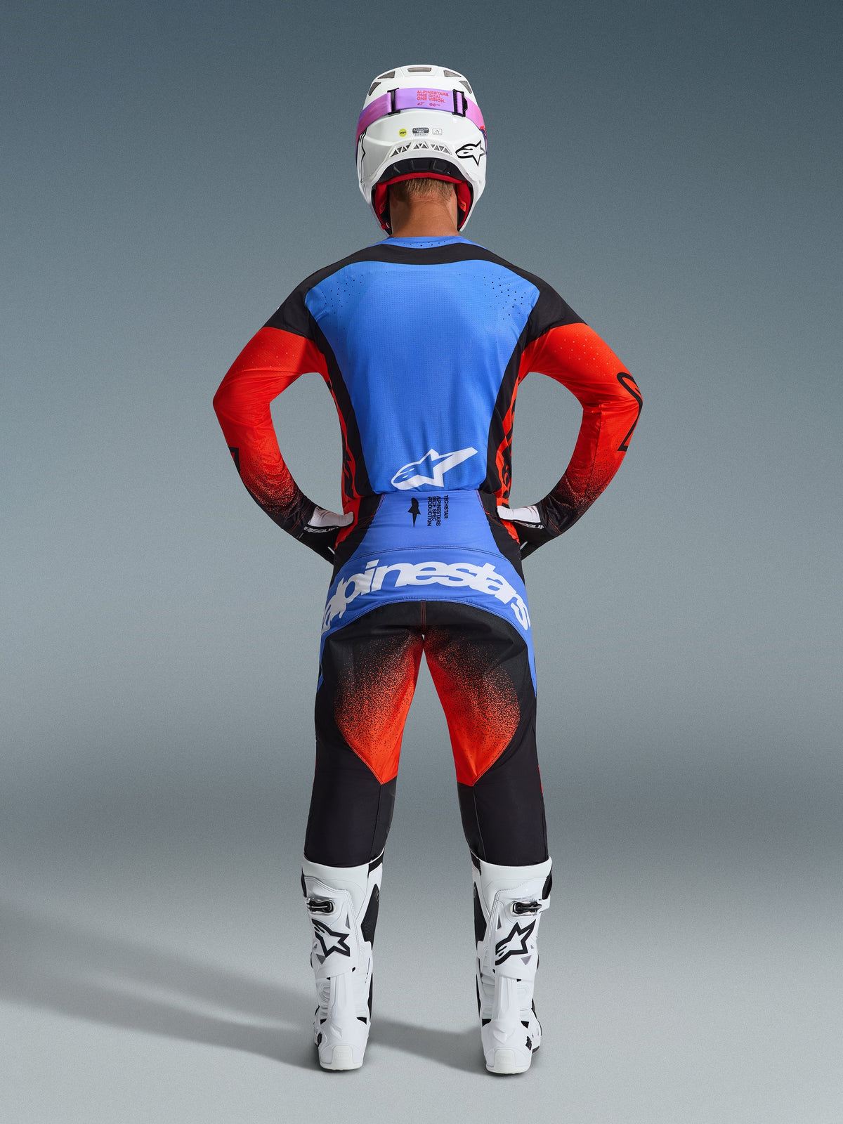 Motocross-kører set bagfra iført komplet racerudstyr, 2026 Techstar Knif-trøje med blå ryg og orange ærmer, sorte bukser med orange og blå detaljer, hvid hjelm med lyserød brillerem, hvide og sorte støvler, koordineret outfit til off-road racing