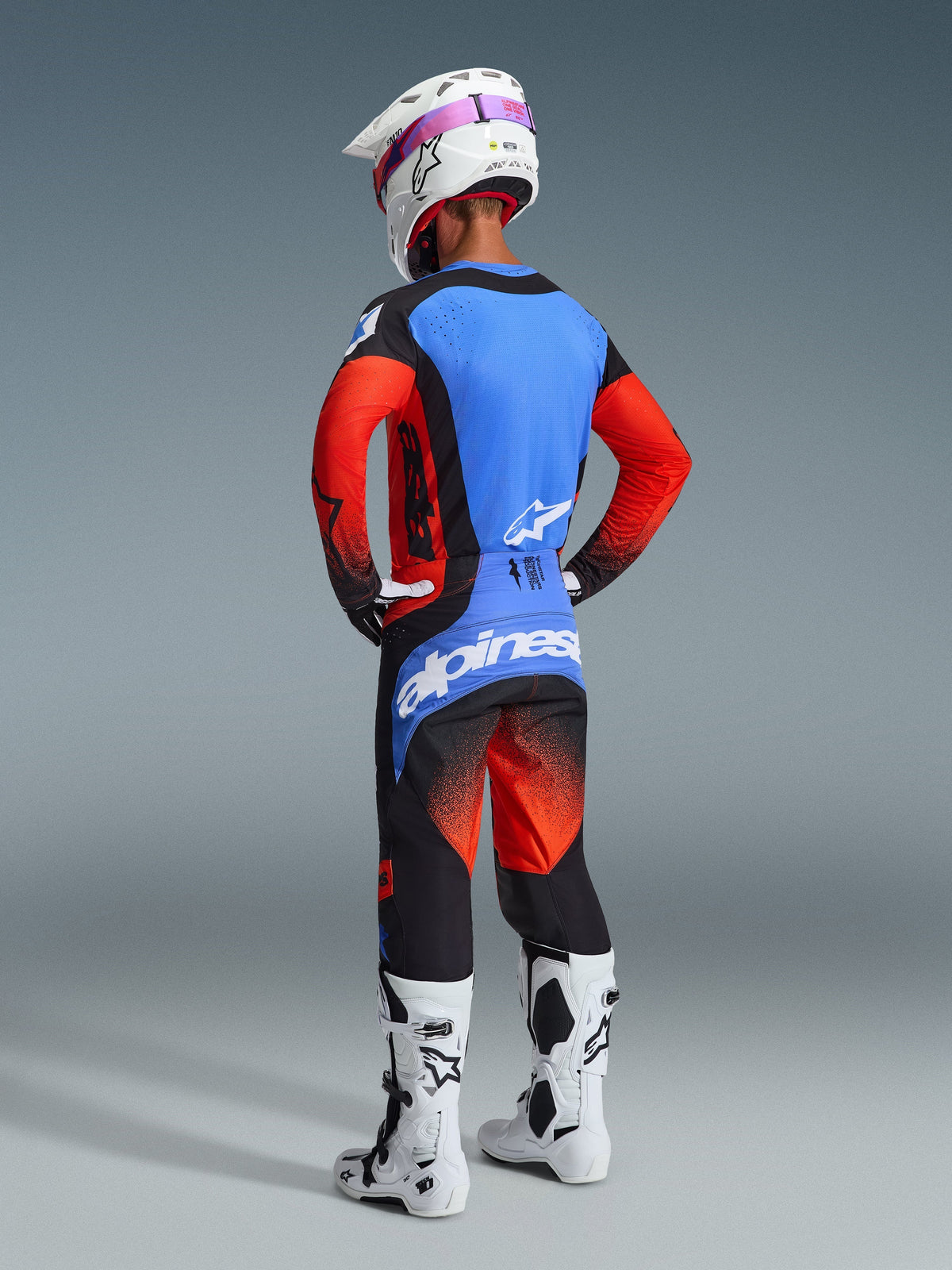 Motocross-kører set bagfra iført 2026 Techstar Knif trøje i orange, sort og UCLA blå, matchende motocross-bukser, hvid hjelm med lyserøde goggles og hvide støvler, komplet Alpinestars off-road raceroutfit