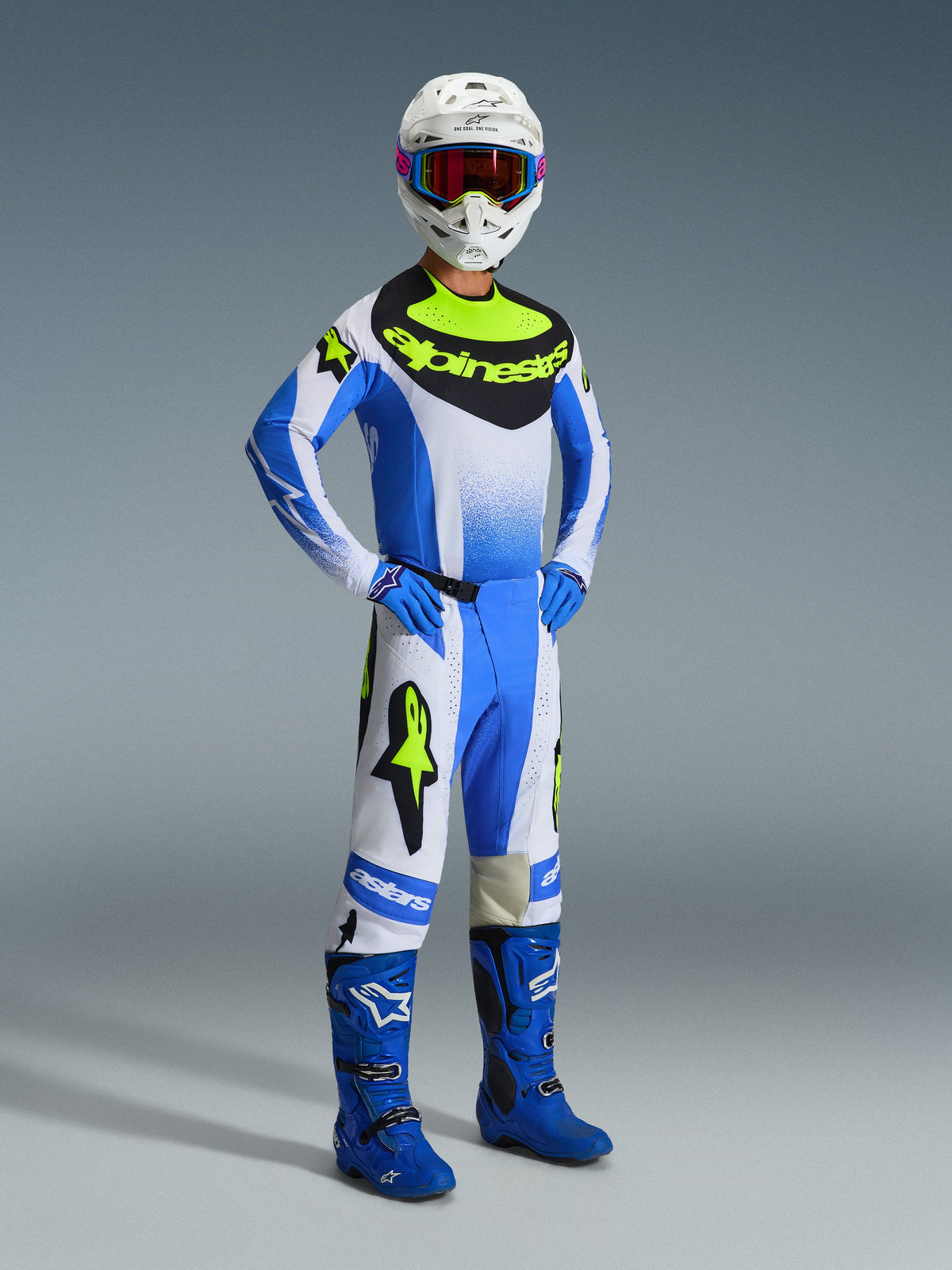 Motocross-kører iført Alpinestars 2026 Techstar Knif-udstyrssæt i Ucla Blue, sort og gul fluo, bestående af en performance-trøje, matchende bukser med markant branding, blå støvler, blå handsker og en hvid hjelm med goggles, stående mod en grå baggrund.