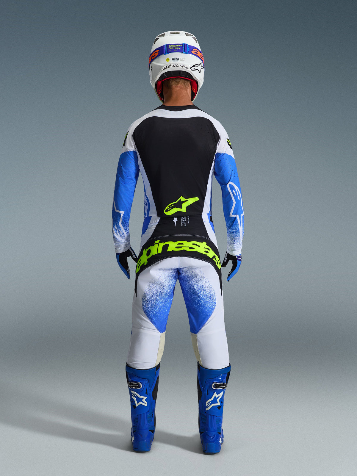 Alpinestars 2026 Techstar Knif-udstyrssæt, set bagfra af en motocross-rytter i Ucla Blue, sort og hvid, med Yellow Fluo-branding på trøjen og bukserne, kombineret med blå racingstøvler og en hvid hjelm.
