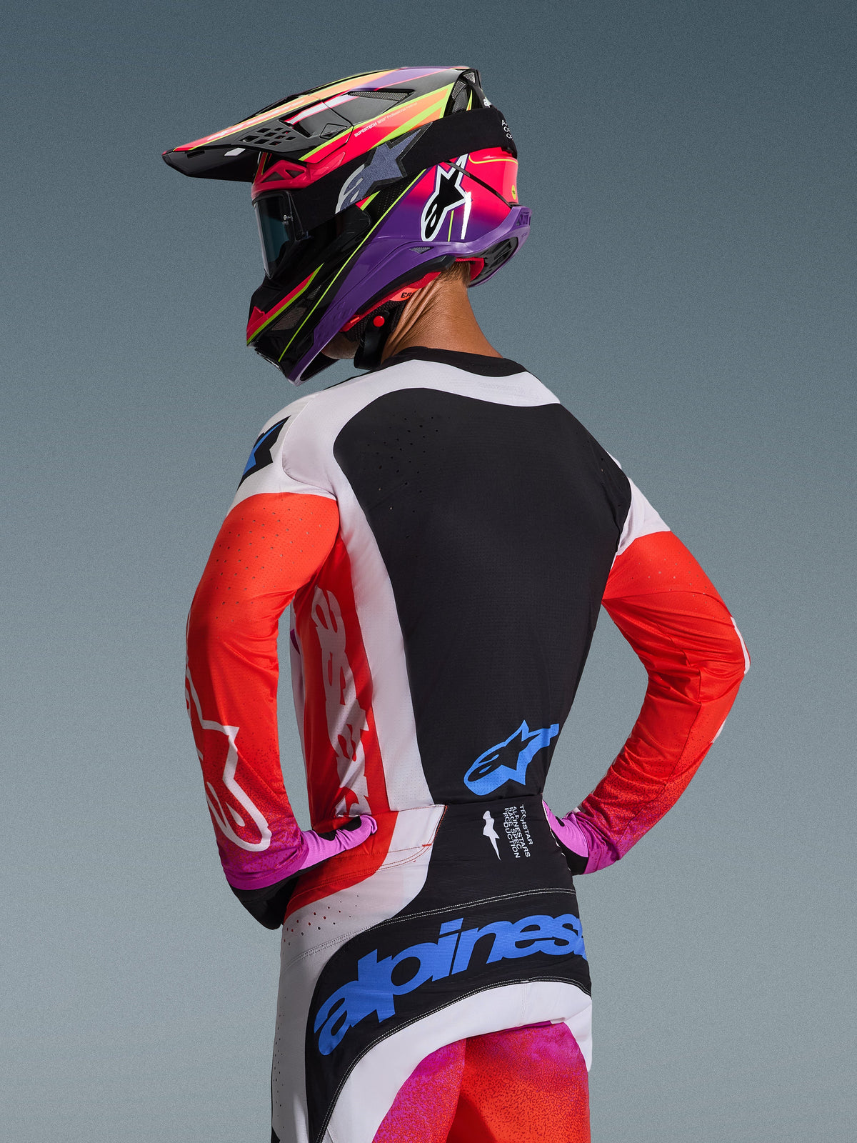 Motocrosskører set bagfra iført Alpinestars 2026 Techstar Knif-udstyrssæt, orange fluo-ærmer med sort rygpanel, hvide og sorte bukser med blå logoer, strålende lilla og pink hjelm, professionelt motocross-racerudstyr til terrænkørsel