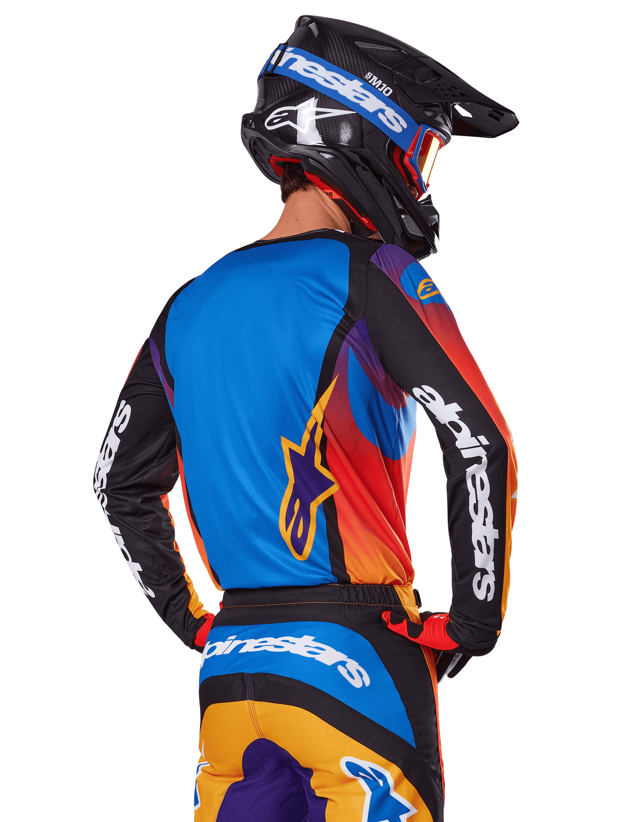 Motocrossrytter set bagfra iført Alpinestars 2025 Fluid Wurx-udstyrssæt, flerfarvet trøje med blå ryg og sorte ærmer med hvide logoer, matchende orange og blå bukser, sort og blå hjelm med orange briller, koordineret off-road raceroutfit.