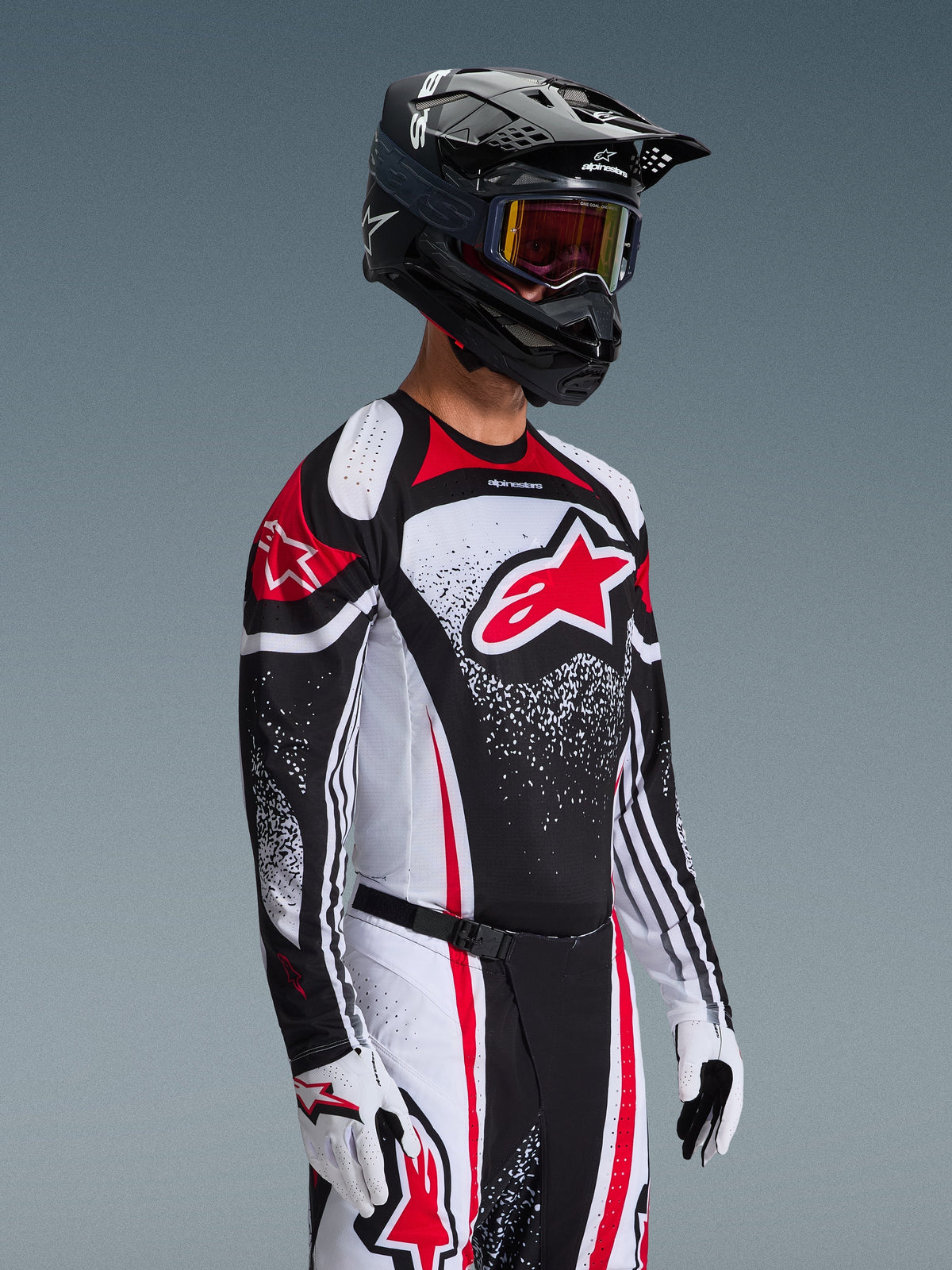 Motocross-kører iført Alpinestars 2026 Techstar Nomur Jersey i sort, hvid og rød, med et stort rødt Astars-logo og nistret grafisk mønster, kombineret med matchende racerbukser, hvide handsker og en sort hjelm med goggles, koordineret outfit til off-road konkurrence