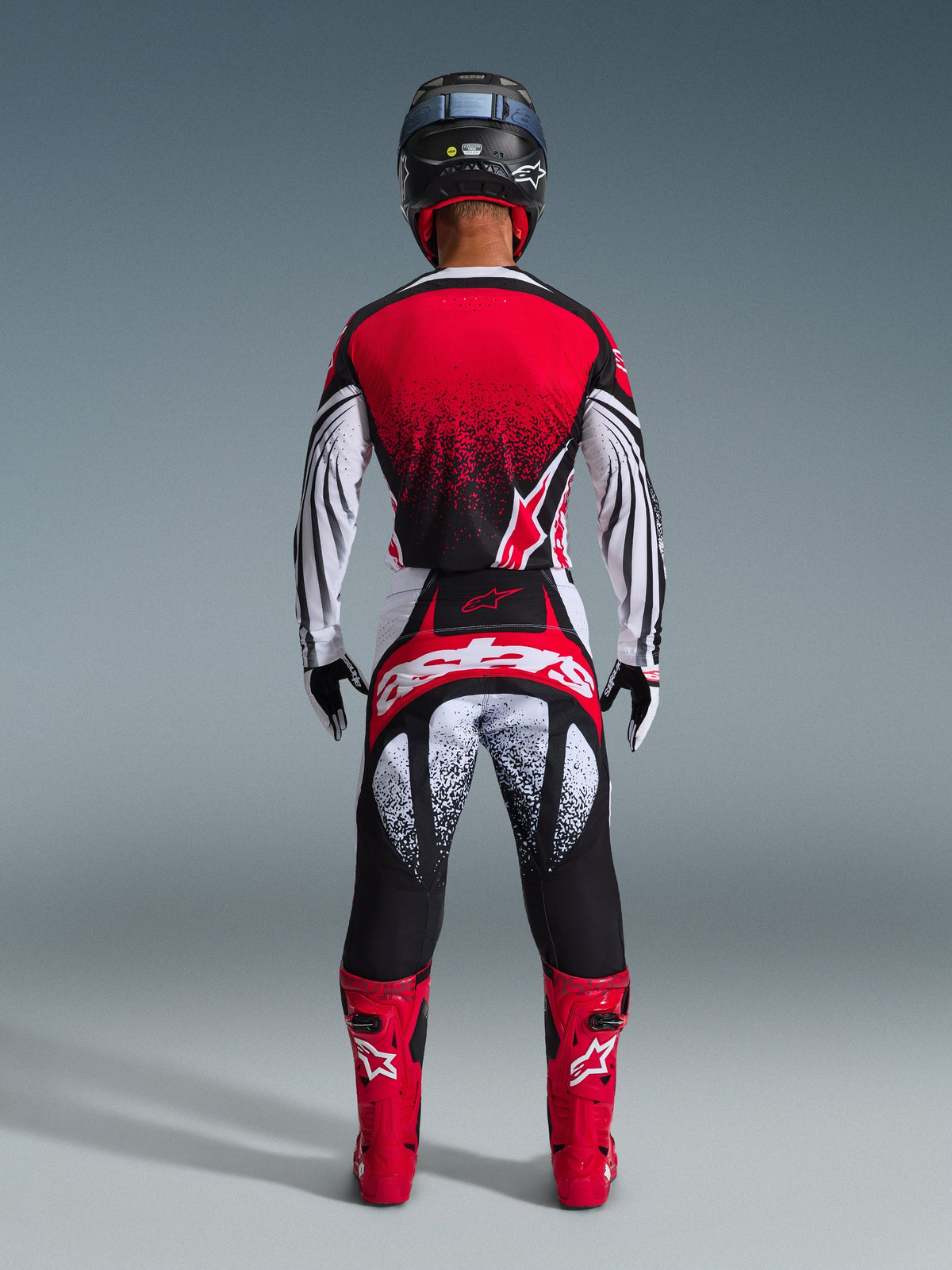 Alpinestars 2026 Techstar Nomur-trøje, motocrossrytter set bagfra, rød, sort og hvid med spættet mønster, matchende racerbukser, røde motocrossstøvler og sort hjelm, komplet udstyrssæt til off-road konkurrence