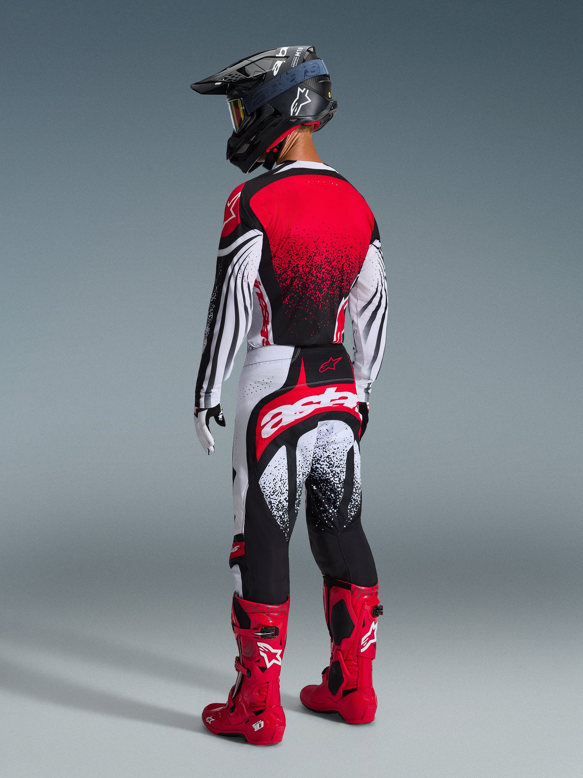 Set bagfra af en motocrosskører iført Alpinestars 2026 Techstar Nomur trøje i sort, hvid og rød, med rød gradient på ryggen og bølgede mønstre på ærmerne, kombineret med matchende motocrossbukser, røde støvler og en sort hjelm for et koordineret off-road racersæt