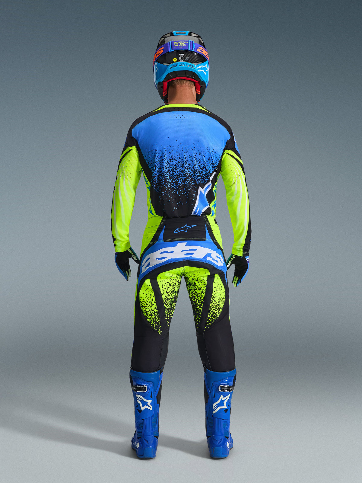 Set bagfra af en motocrosskører iført Alpinestars 2026 Techstar Nomur-udstyrssæt. Sættet inkluderer en blå og neongul jersey med sorte spraymønster-accenter, matchende neongule og sorte bukser med et stort blåt 'astars'-logo på sædet, koordinerede blå støvler med hvide logoer og en blå og sort hjelm. Komplet professionelt racerudstyr designet til ydeevne og synlighed.