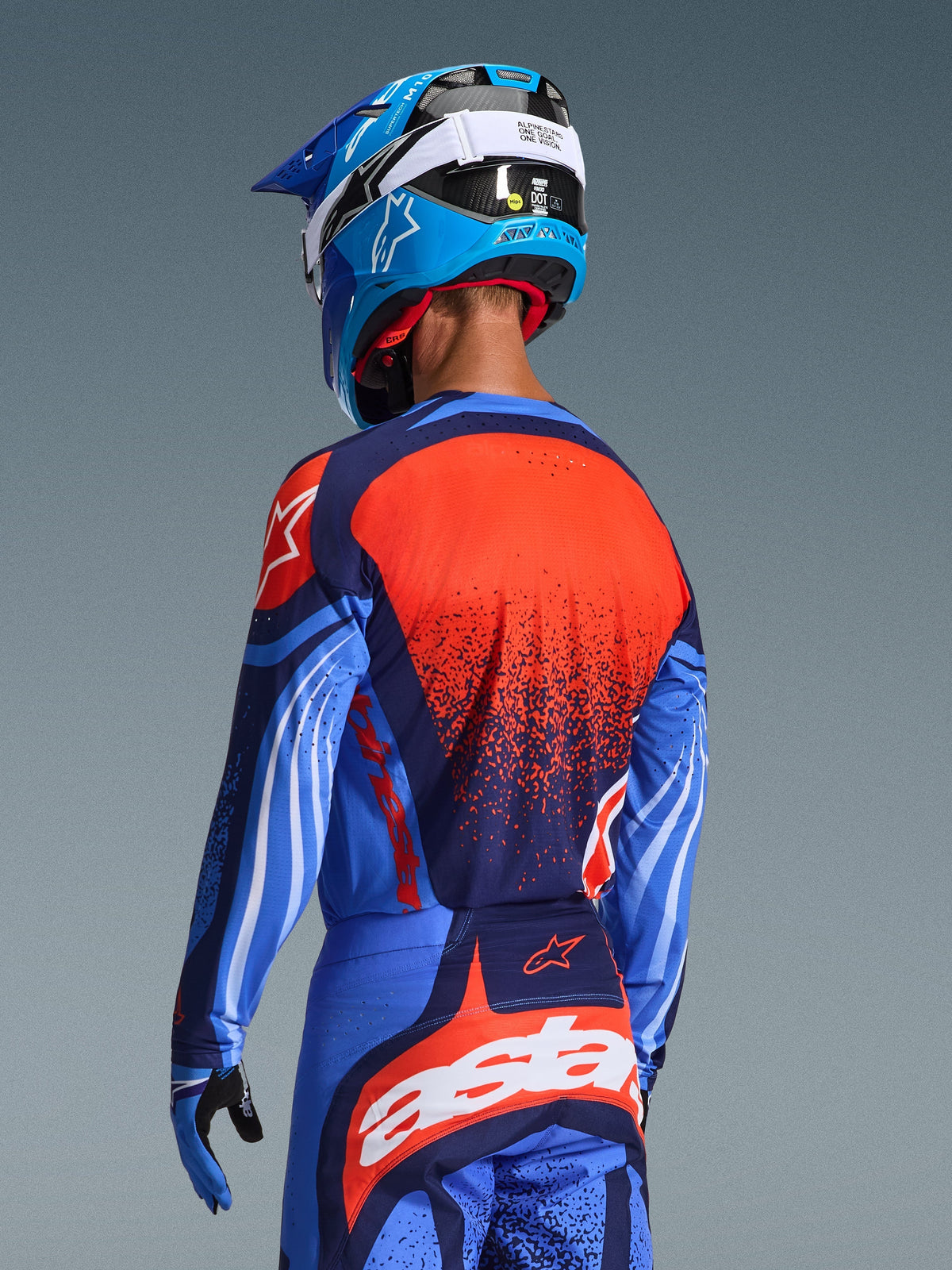 Motocross-rytter set bagfra iført Alpinestars 2026 Techstar Nomur-trøje og matchende bukser, mørkeblå og UCLA-blå med livlige orange detaljer, med en stor stjerne-grafik og laserskåret ventilation på trøjen, kombineret med en blå og hvid hjelm for et komplet off-road racersæt