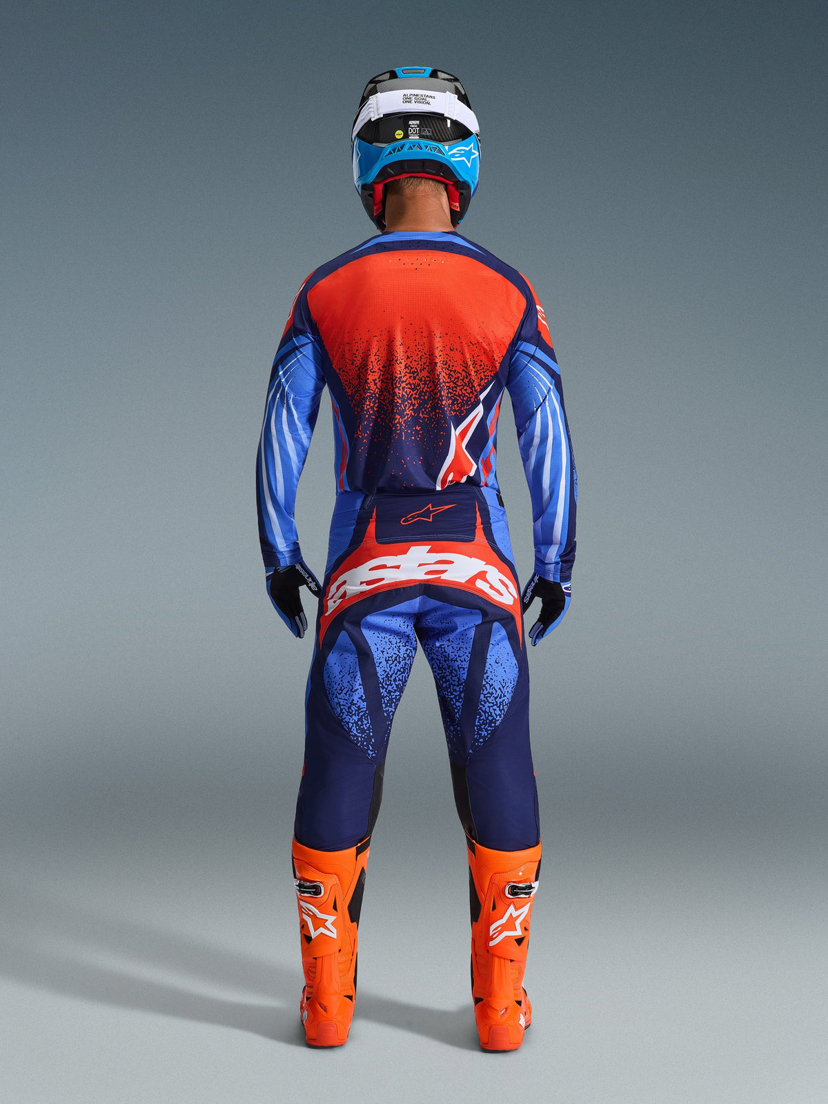 Motocross-kører set bagfra iført 2026 Techstar Nomur-trøje og matchende bukser, mørkeblå og orange farvekombination med UCLA-blå detaljer, stærkt orange støvler, blå og hvid hjelm og sorte handsker, komplet professionelt off-road racingudstyr