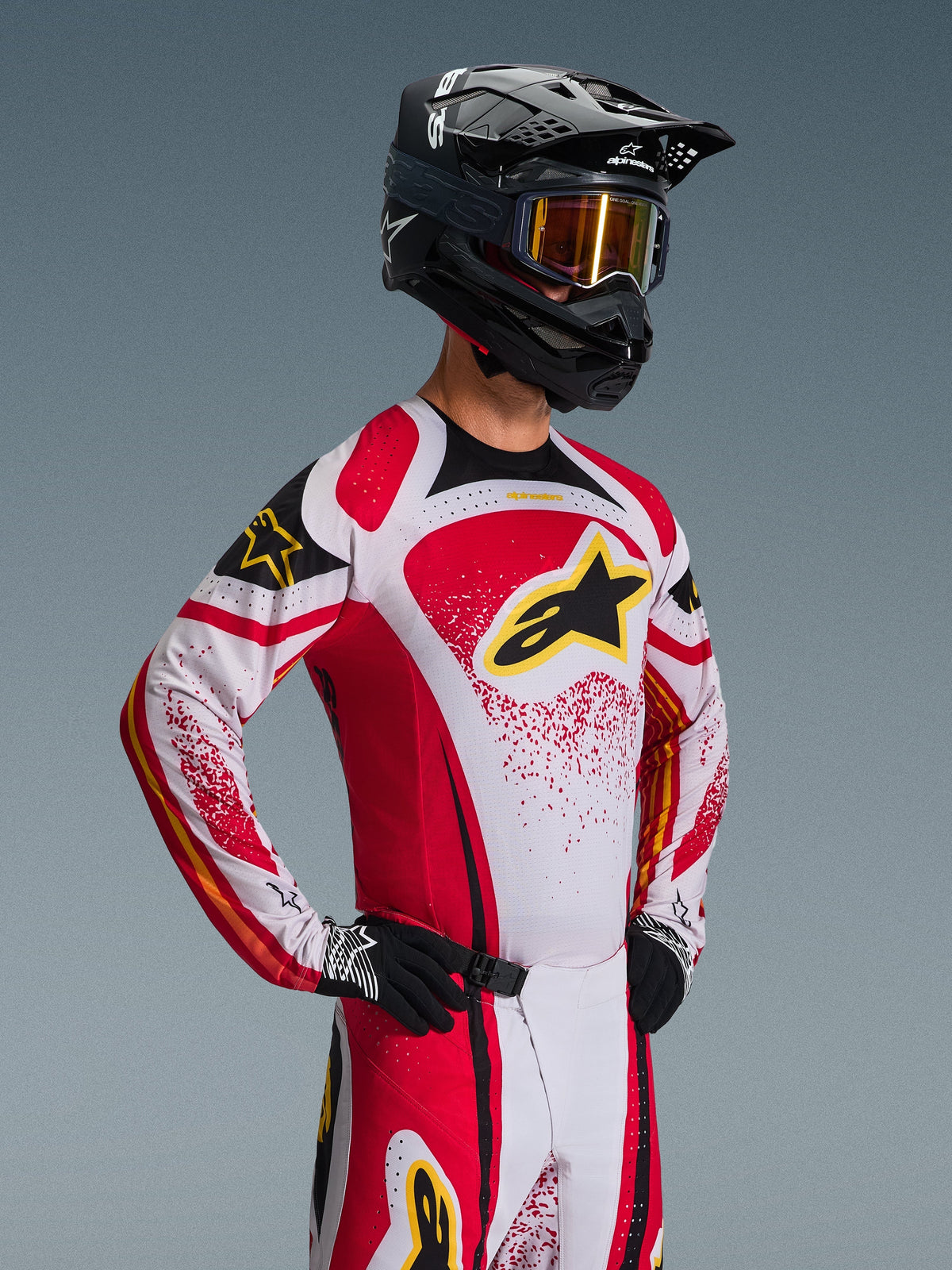 Motocross-rytter iført Alpinestars 2026 Techstar Nomur Jersey, rød, hvid og sort med gule detaljer, kombineret med matchende bukser, sort hjelm med guld-goggles og sorte handsker, atletisk race-pasform til professionel off-road-konkurrence