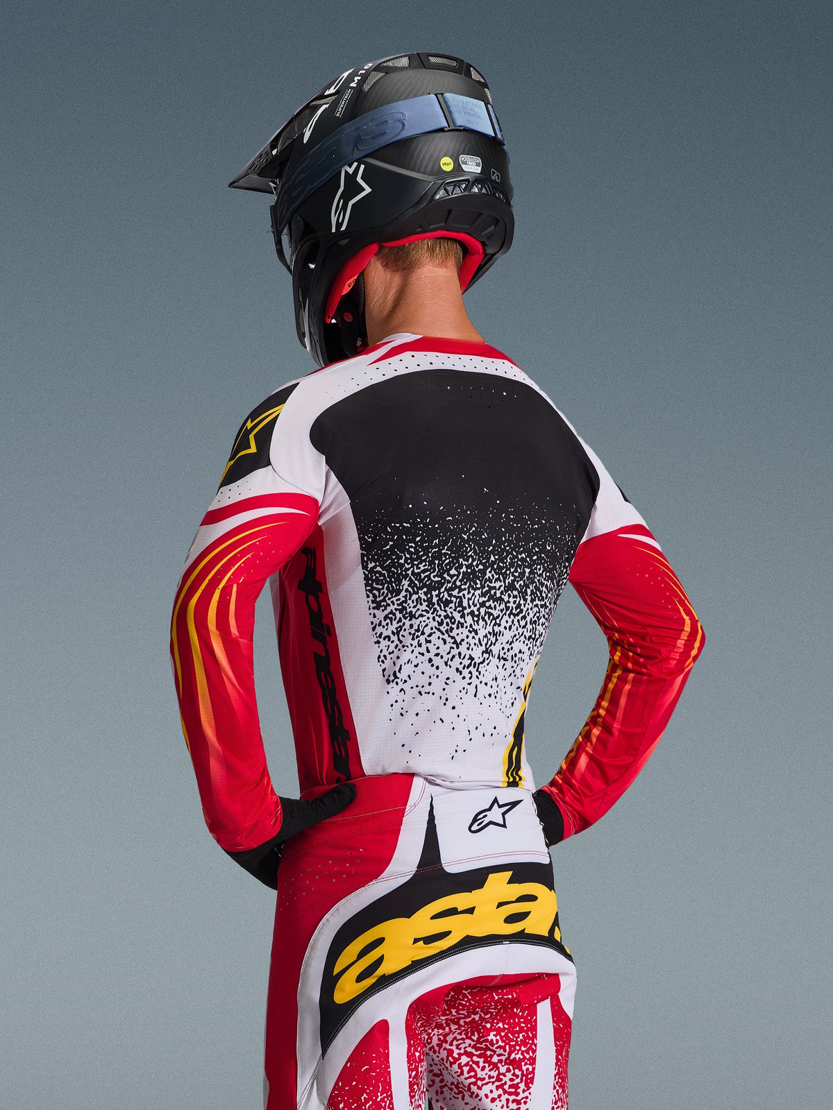 Motocrosskører set bagfra iført Alpinestars 2026 Techstar Nomur trøje og matchende bukser, rød, hvid og sort med gule detaljer, med et nisteret gradientdesign på ryggen, sort Alpinestars hjelm og sorte handsker, koordineret professionelt udstyr til off-road racing