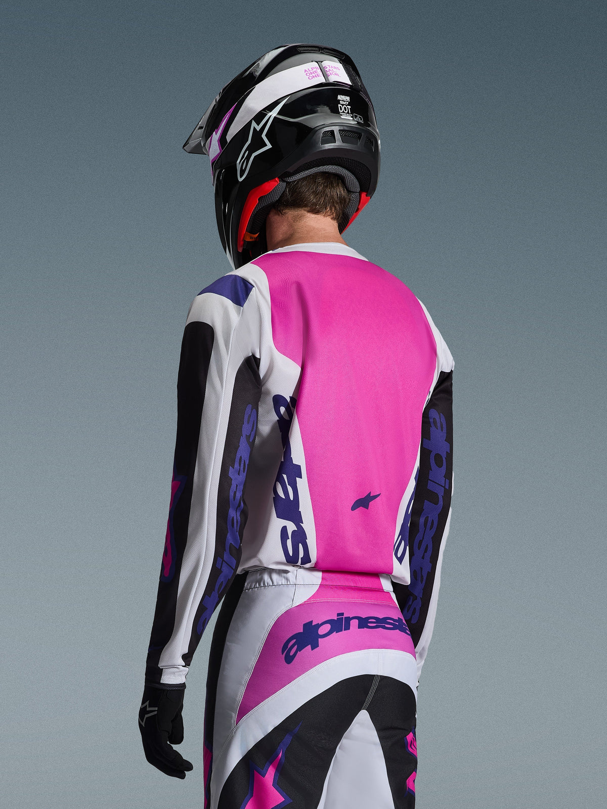 Motocrosskører set bagfra iført Alpinestars 2026 Fluid Grid trøje i sort, hvid og lilla, matchende bukser med markant branding, sort hjelm med lyserøde detaljer og sorte handsker, komplet koordineret udstyr til off-road kørsel.