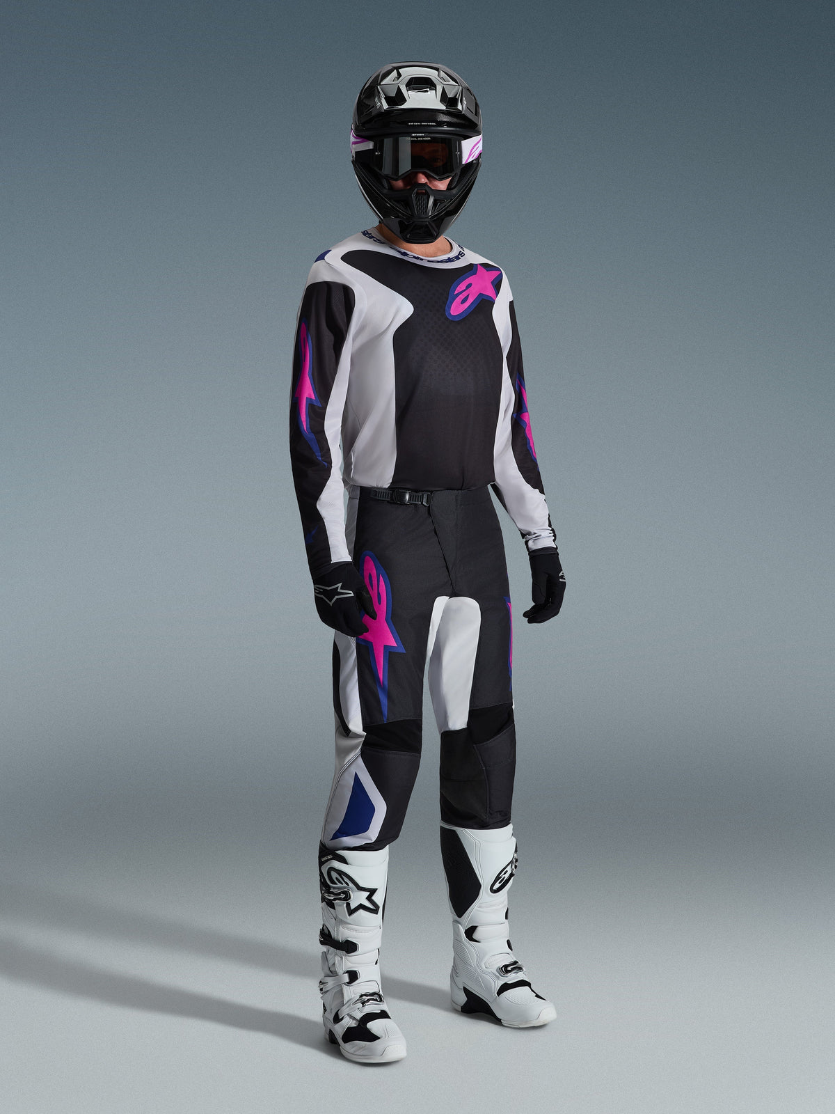 Motocross-kører iført komplet racerudstyr, 2026 Fluid Grid-trøje i sort med lysegrå og lilla detaljer, matchende Fluid Grid-bukser, sort hjelm med koordinerede goggles, hvide og sorte støvler og sorte handsker, professionel studieopsætning til off-road racing.