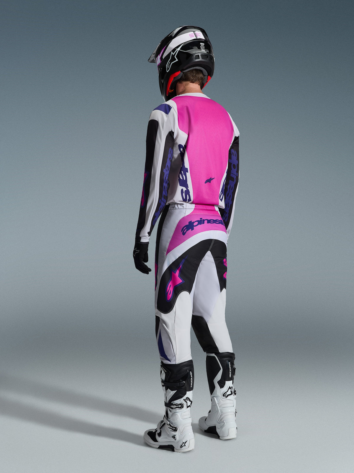 Motocross-rytter set bagfra iført Alpinestars 2026 Fluid Grid trøje og matchende bukser, lysegrå og sort med et stærkt lyserødt rygpanel og lilla branding, fuldendt med en sort hjelm og hvide støvler.