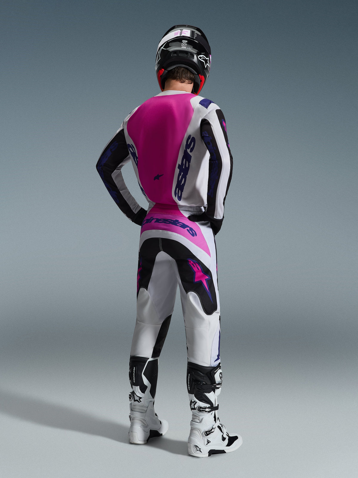 Motocross-kører set bagfra iført Alpinestars 2026 Fluid Grid trøje, hvid trøje med stort pink rygpanel og lilla branding, matchende hvide og sorte bukser med pink detaljer, hvide støvler og sort hjelm, koordineret off-road racerudstyr