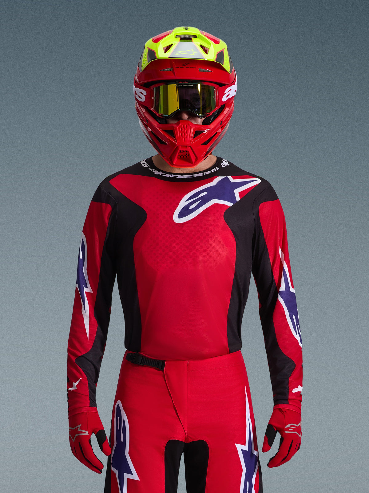 Motocross rider wearing Alpinestars 2026 Fluid Grid Gear Set, Red jersey with black side panels and white star logo, matching red pants and gloves, red and yellow helmet with gold tinted goggles, high-performance racing gear for off-road
