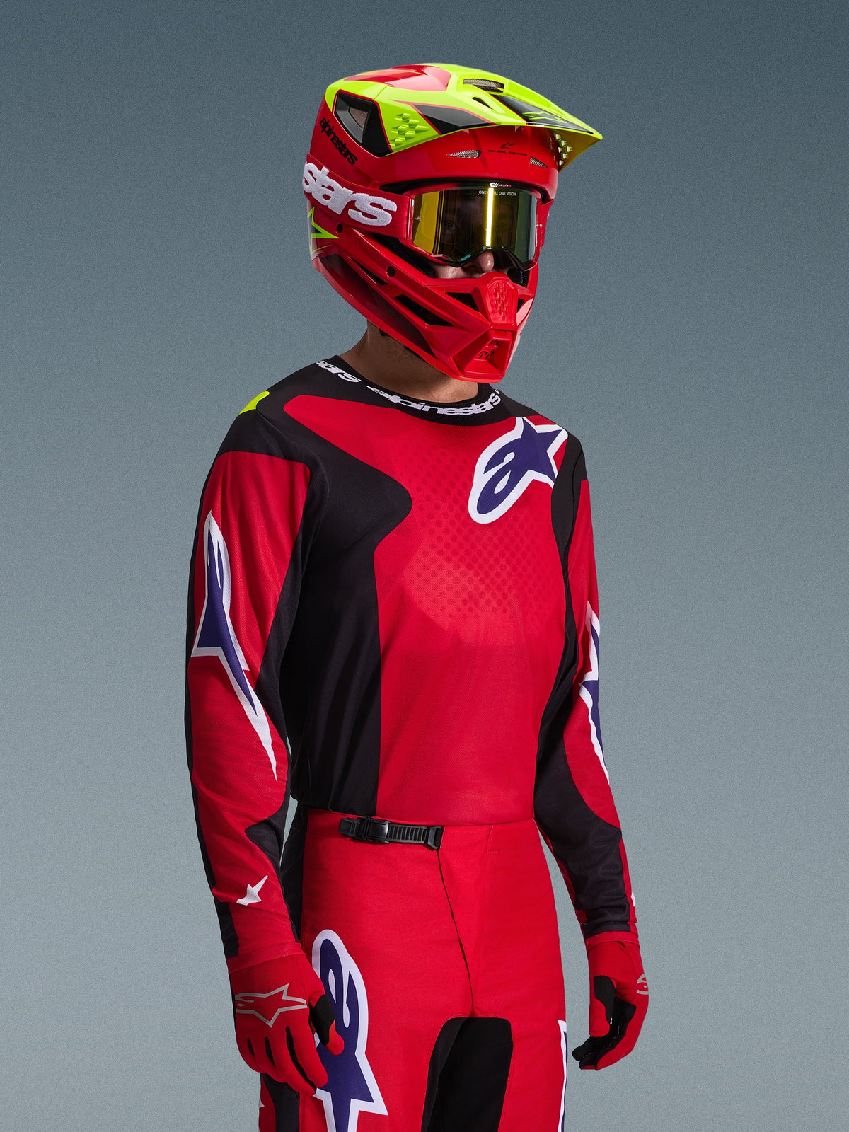 Motocross-kører iført Alpinestars 2026 Fluid Grid-trøje i rød, sort og lilla, koordineret med matchende motocross-bukser, en rød hjelm med neongule detaljer, guld-goggles og røde handsker, komplet off-road raceroutfit.