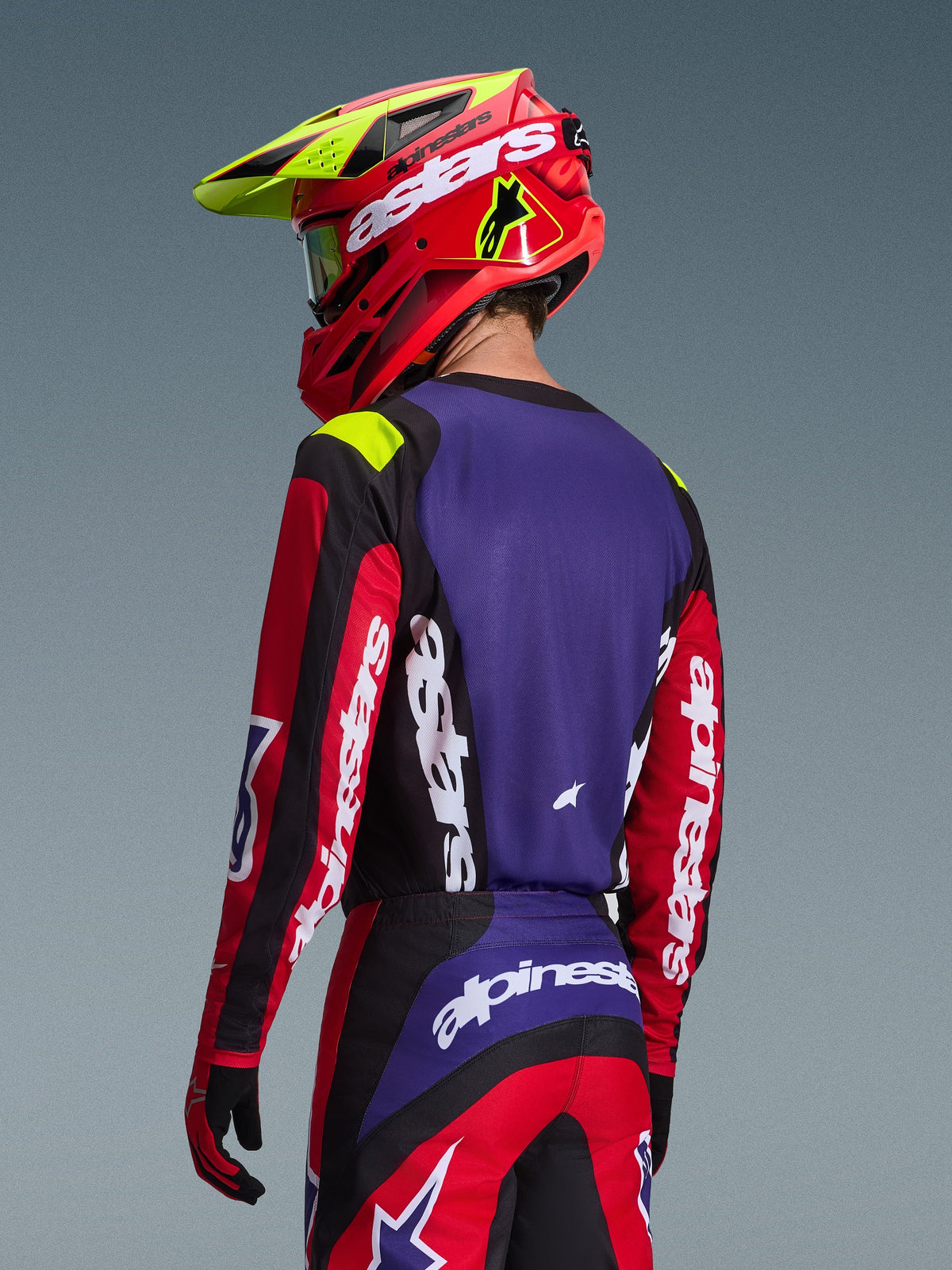 Motocross-kører iført Alpinestars 2026 Fluid Grid trøje, rød, sort og lilla, set bagfra med lilla rygpanel og røde ærmer med hvidt branding, matchende bukser og en rød og neongul hjelm, koordineret sæt til off-road racing