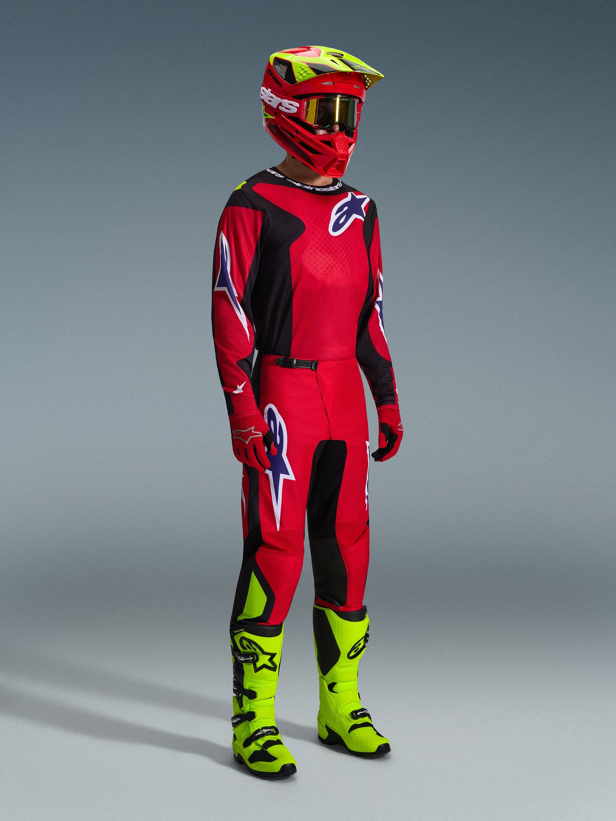 Alpinestars motocross-kører i komplet racerudstyr, 2026 Fluid Grid-trøje i rød, sort og lilla, matchende bukser med lilla logo-detaljer, rød og neongul hjelm med guldbriller og neongule støvler, koordineret outfit til off-road racing