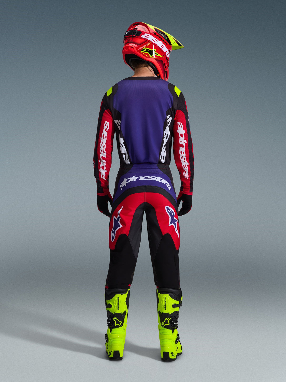 Motocross rider from behind wearing the Alpinestars 2026 Fluid Grid Gear Set in Red Black Purple, featuring a purple back panel and red sleeves on the jersey, matching red and black pants, bright neon yellow motocross boots, and a red and neon yellow helmet.