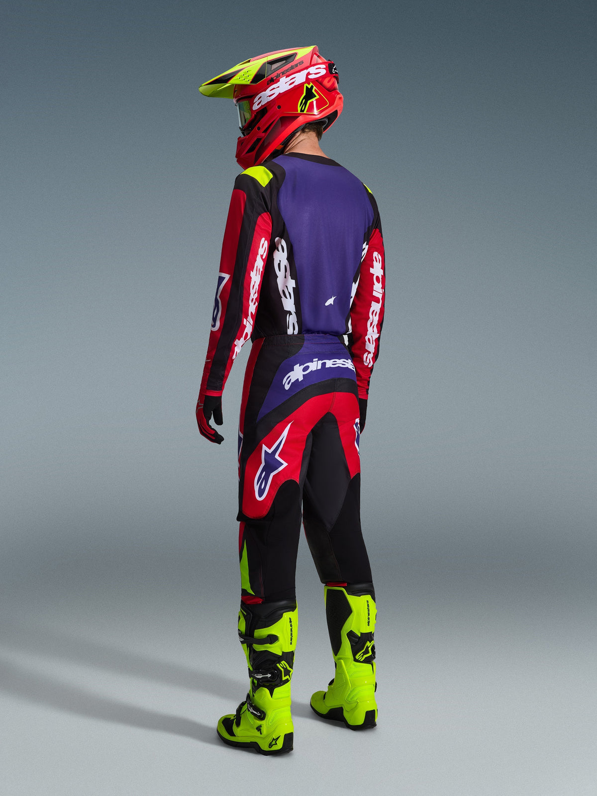 Motocross rider wearing the Alpinestars 2026 Fluid Grid Gear Set from the back, Red Black and Purple jersey with white branding, red and black pants with purple highlights, coordinated with a red and neon yellow helmet and bright neon yellow boots, full off-road racing gear.