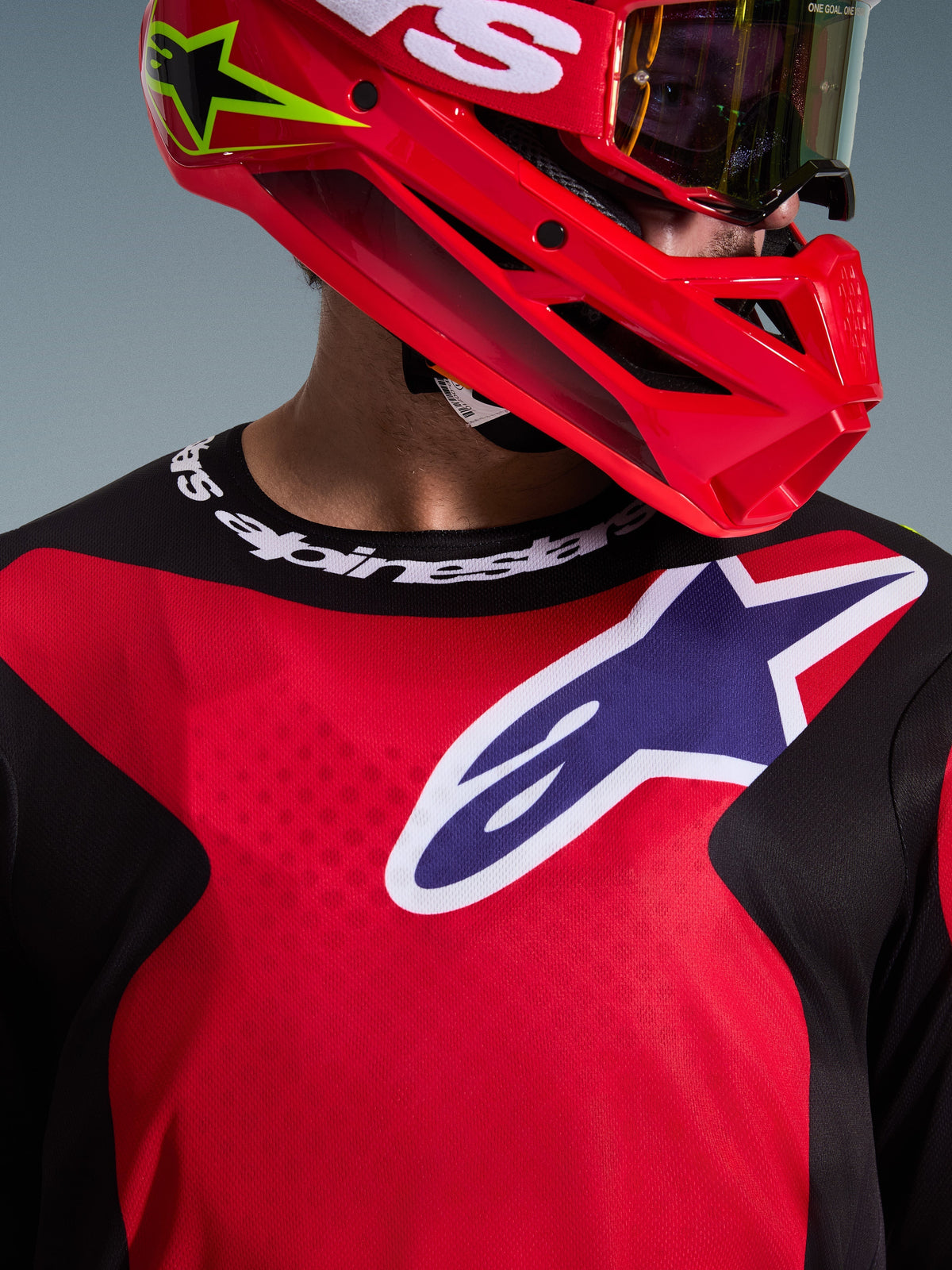 Nærbillede af en motocrosskører iført 2026 Fluid Grid Jersey i rød, sort og lilla, med et stort Alpinestars-logo på brystet og sportskrave med logo, kombineret med en rød hjelm og goggles, der fremhæver det funktionelle poly-tekstil og den atletiske pasform.