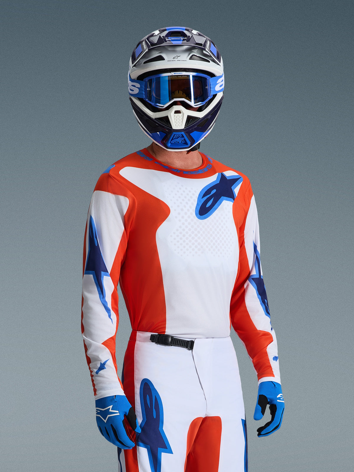 Motocross-kører iført Alpinestars 2026 Fluid Grid-udstyrssæt, orange og hvid jersey med blå stjernelogoer, hvide og orange bukser med blå detaljer, blå handsker, blå og hvid hjelm med blå goggles, koordineret atletisk racerudstyr til off-road-performance