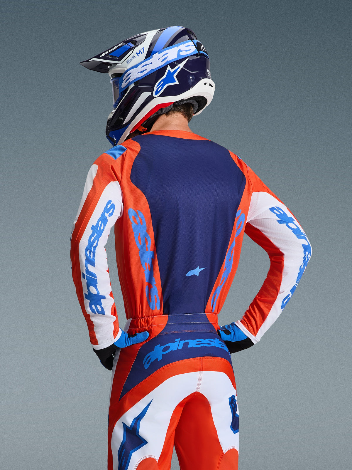 Alpinestars 2026 Fluid Grid Jersey, Motocross Jersey, Orange and Ucla Blue, rear view of rider wearing matching pants and helmet, featuring navy blue back panel and white sleeves with light blue branding, athletic fit for off-road racing