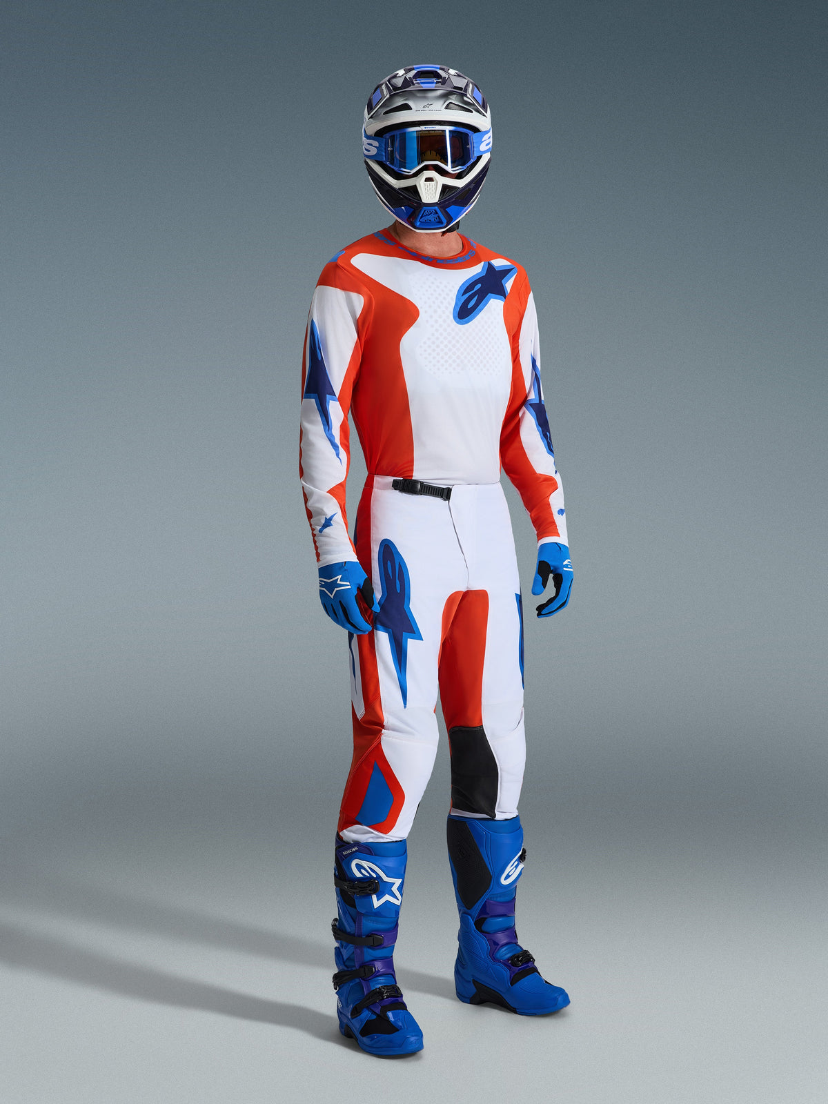 Alpinestars 2026 Fluid Grid-udstyrssæt, motocross-kører med hvid trøje og bukser, orange og UCLA-blå grafik, matchende blå hjelm og briller, blå handsker og lyseblå racestøvler, koordineret præstationsoutfit til off-road-kørsel
