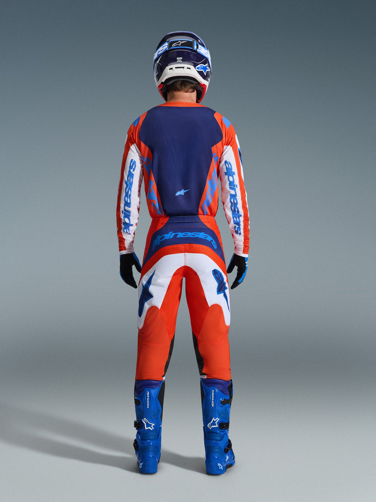 Alpinestars 2026 Fluid Grid Jersey, Motocross Jersey, Orange and UCLA Blue, back view of rider in full racing gear set, featuring a dark blue back panel and white sleeves with blue branding, paired with matching pants and blue boots.