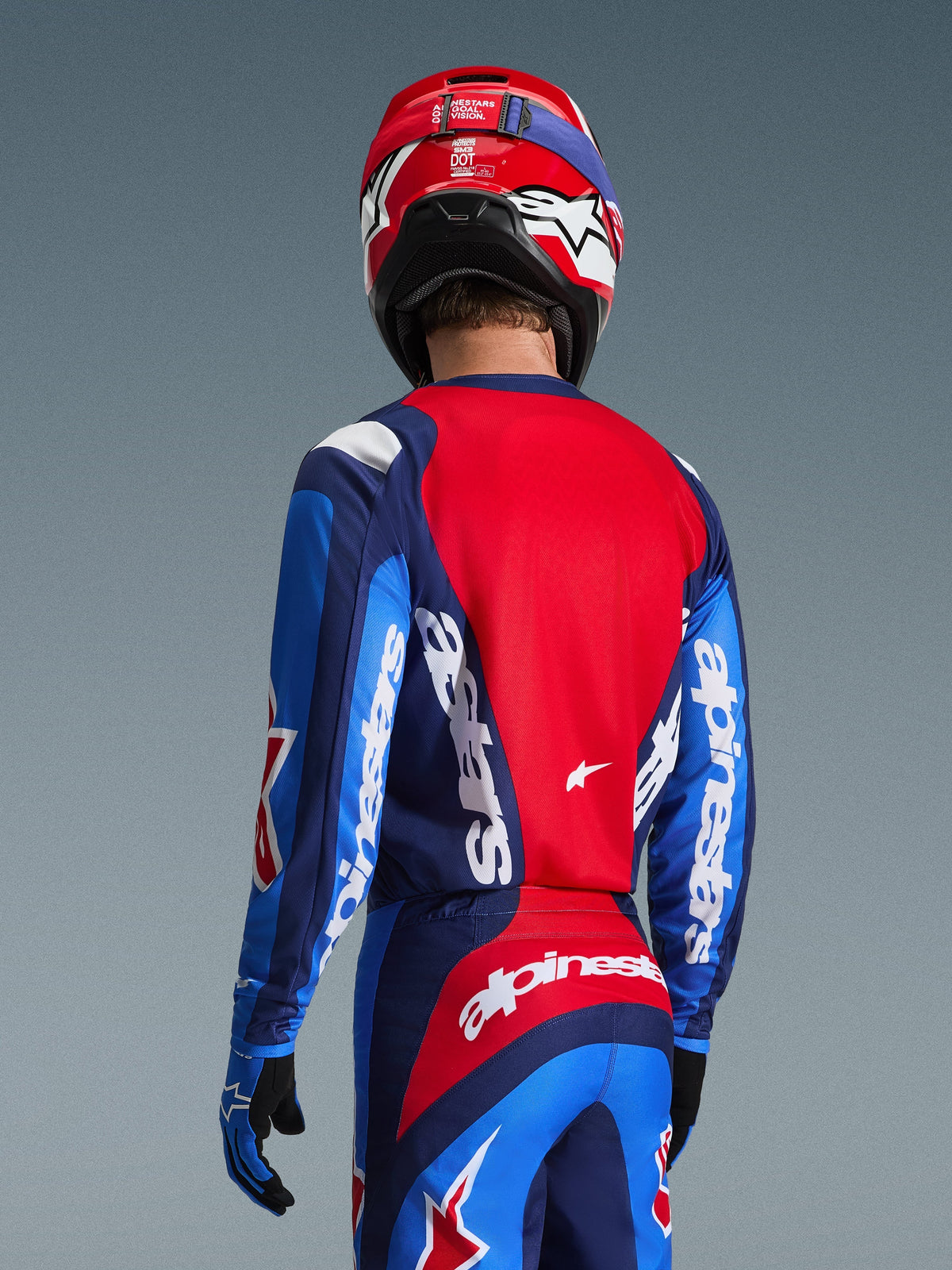 Set bagfra af en motocross-kører iført Alpinestars 2026 Fluid Grid-trøjen i blå og rød, med markant hvid branding på ærmerne og ryggen, kombineret med matchende motocross-bukser og en rød hjelm for et komplet off-road racerlook