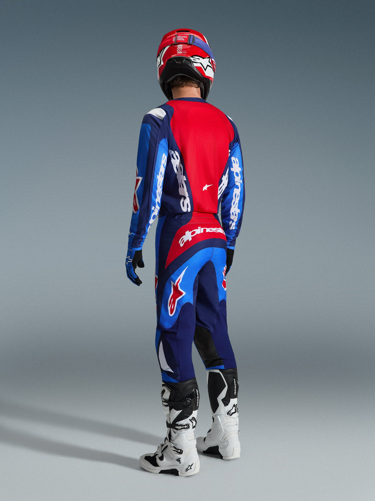 Motocross rider from the rear wearing the Alpinestars 2026 Fluid Grid Jersey in Blue and Red, matching motocross pants with bold white branding, red helmet, and white and black boots, complete off-road racing gear set